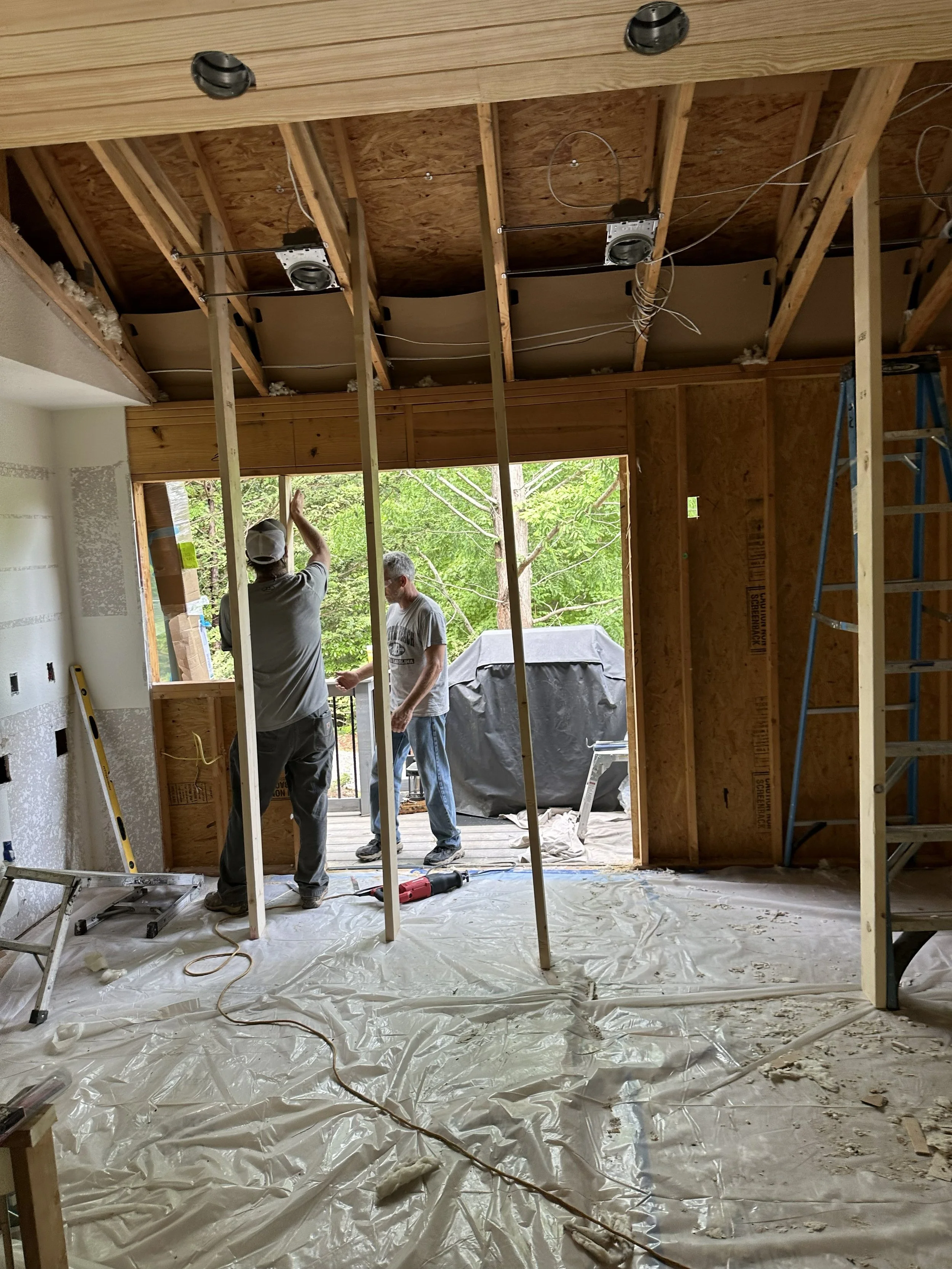 Two construction workers standing inside a partially built room, with exposed wooden framing, wires, and construction tools, during a residential remodeling project.