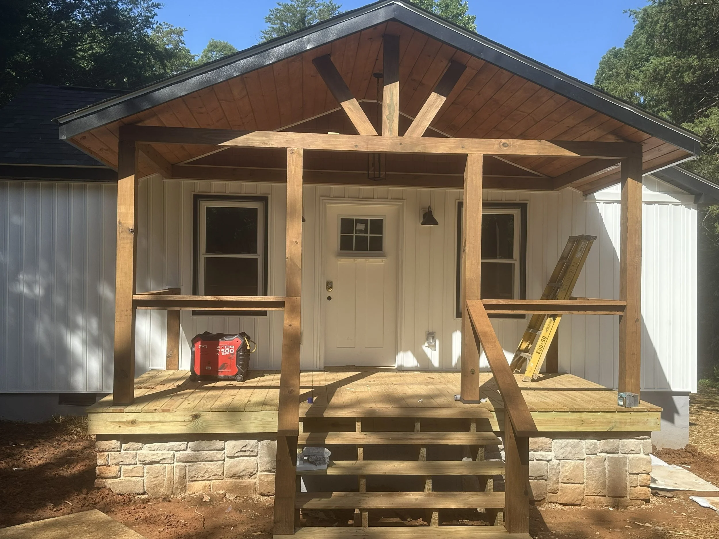 New wooden porch under construction in front of a house with white siding and a stone foundation, with construction tools and a ladder nearby.