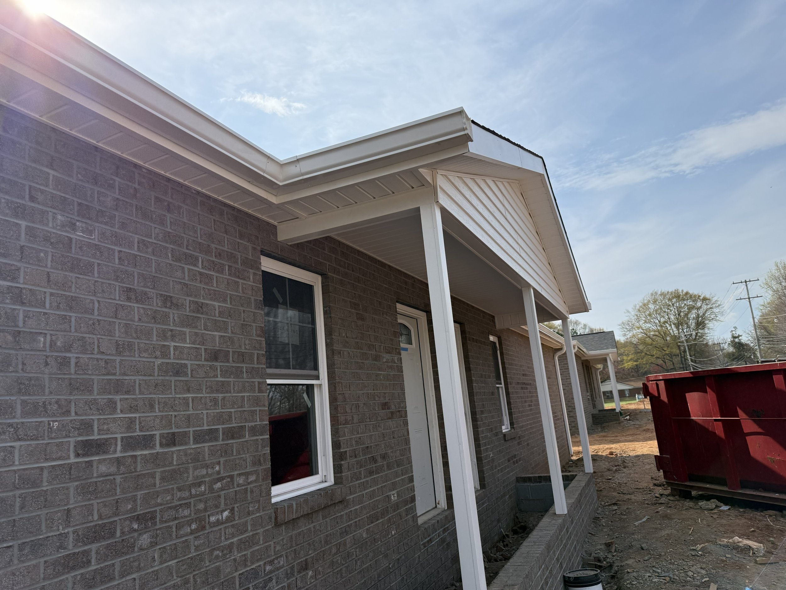 The exterior of a house under construction, showing a brick wall, white siding, and a roof with a gutter, with a partly cloudy sky in the background.