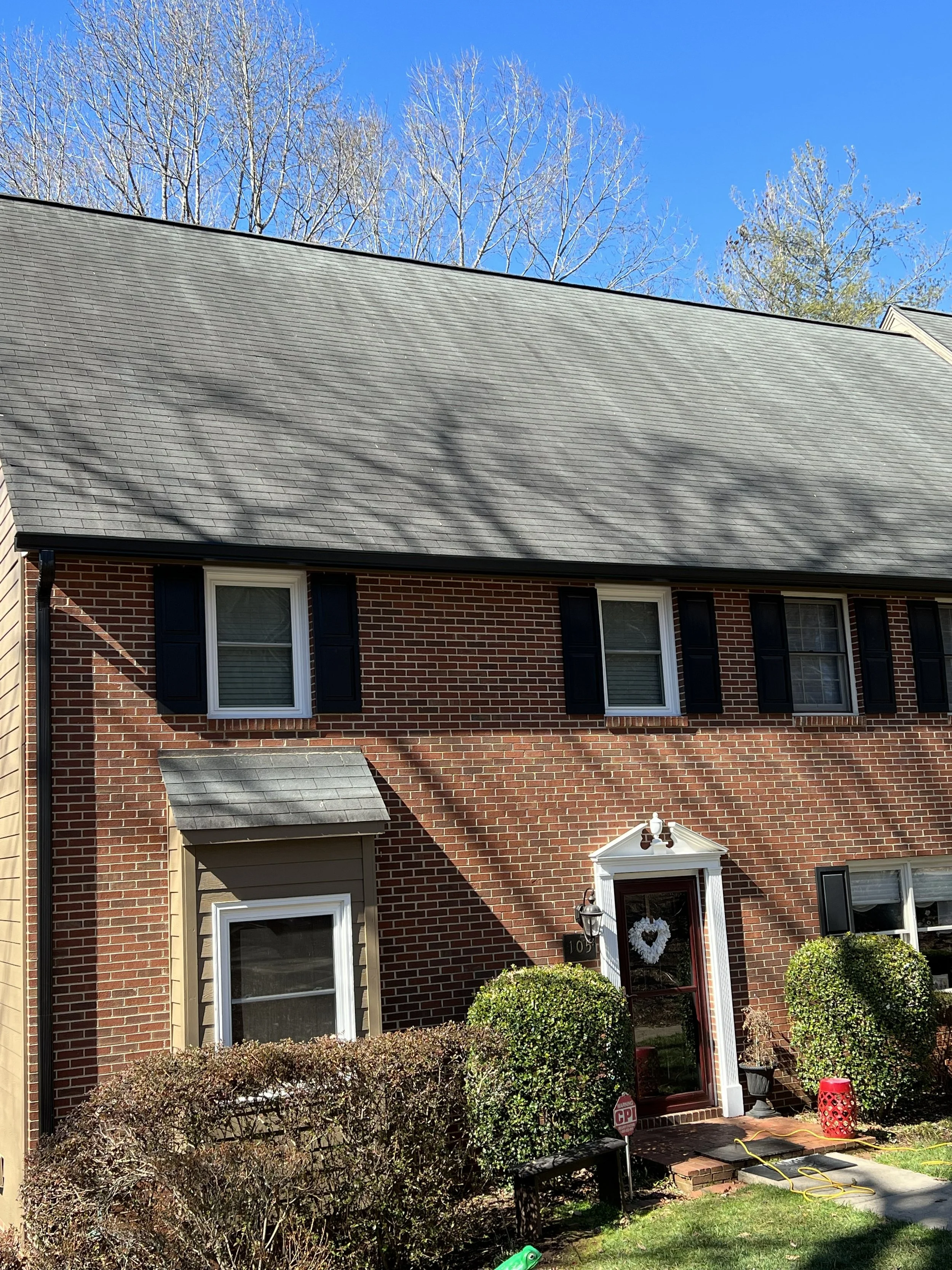 A brick house with three windows on the second floor, black shutters, and a white front door adorned with a heart-shaped wreath. There are bushes in front, and a small porch with indoor lighting. The house has a black roof, and there are leafless tre