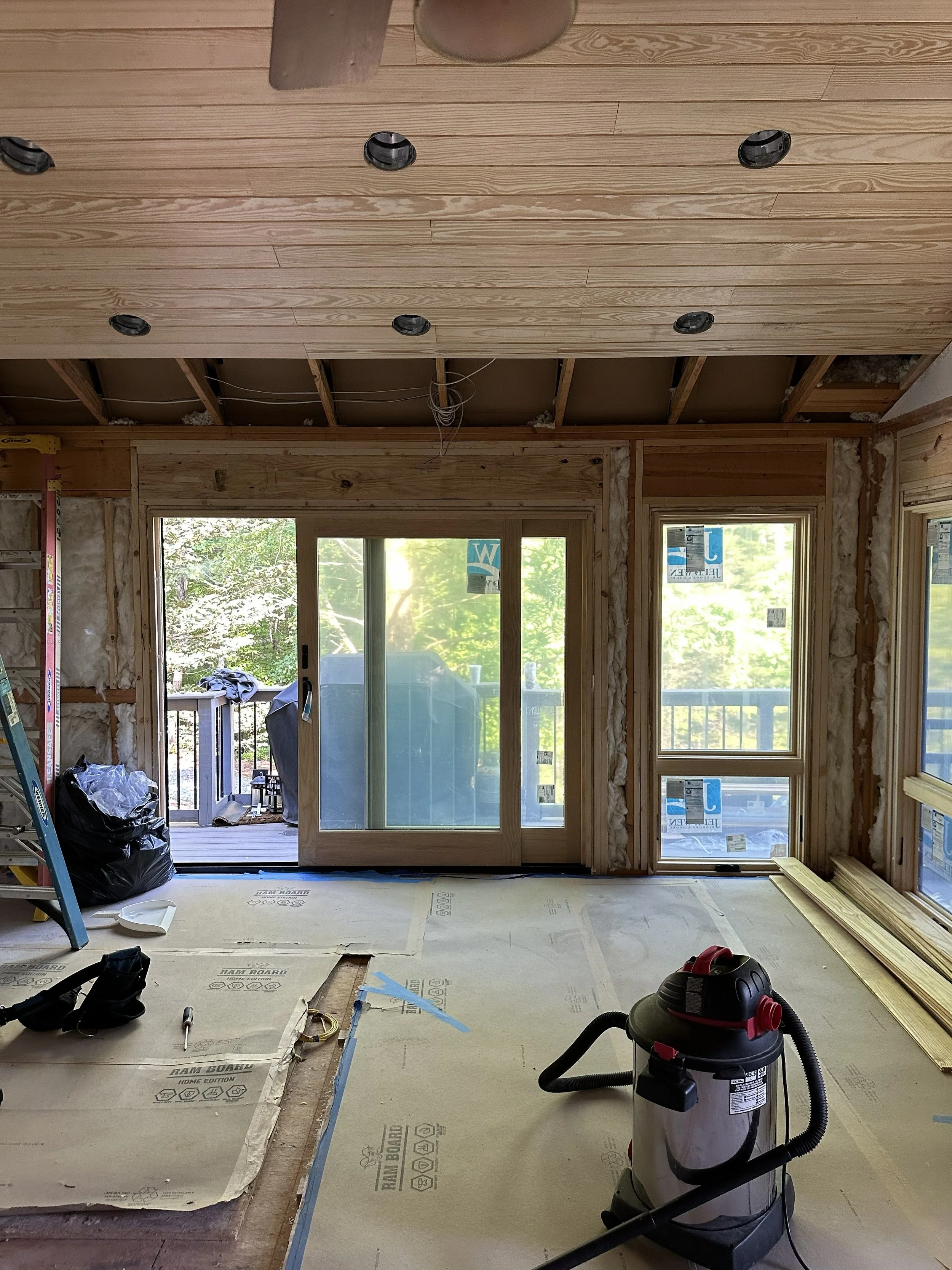 Room under renovation with new wooden sliding doors installed, construction tools, and insulation visible, overlooking a balcony and outside greenery.