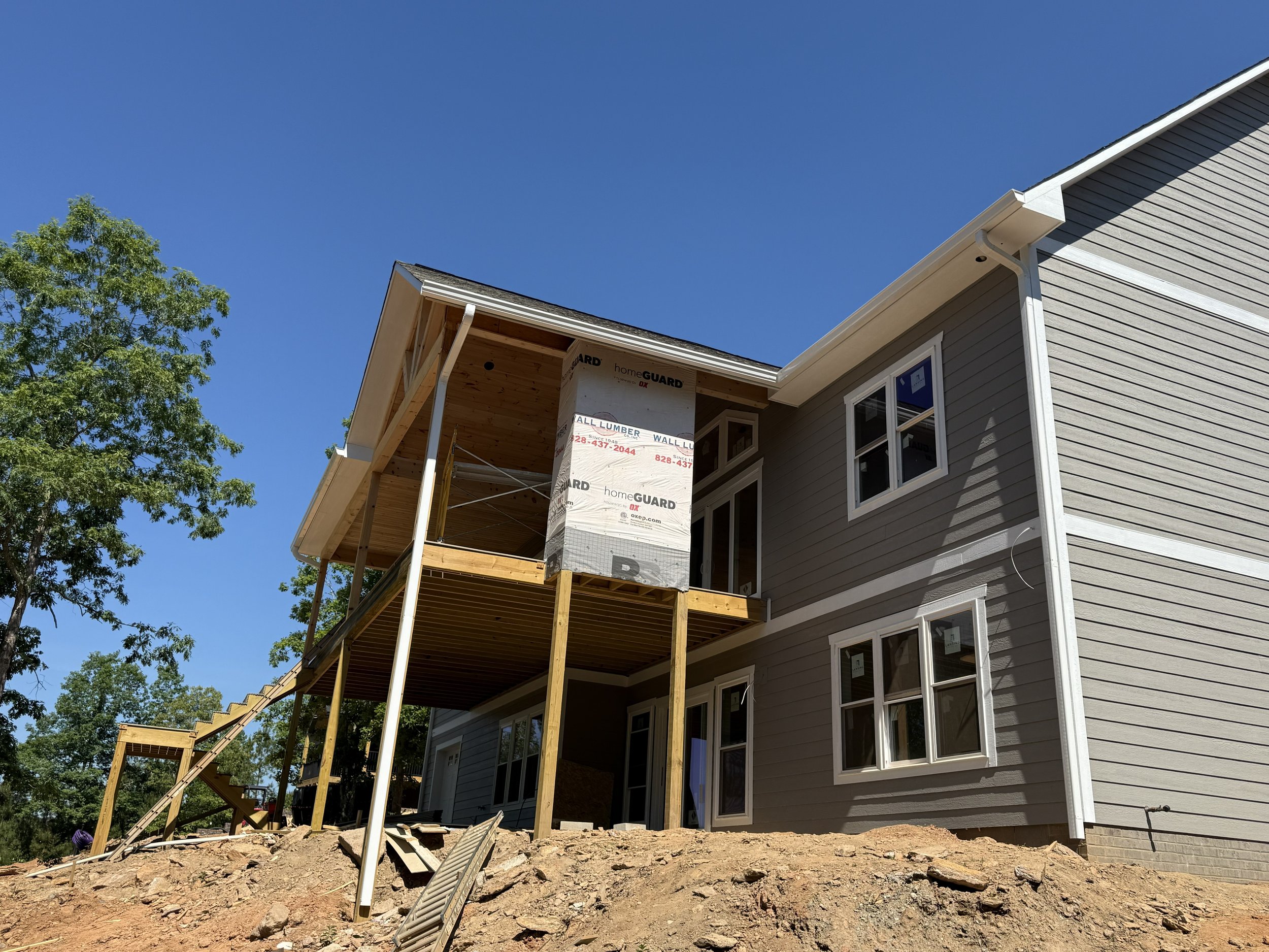 Partially constructed house with scaffolding and wooden stairs, surrounding soil, and trees against a clear blue sky.