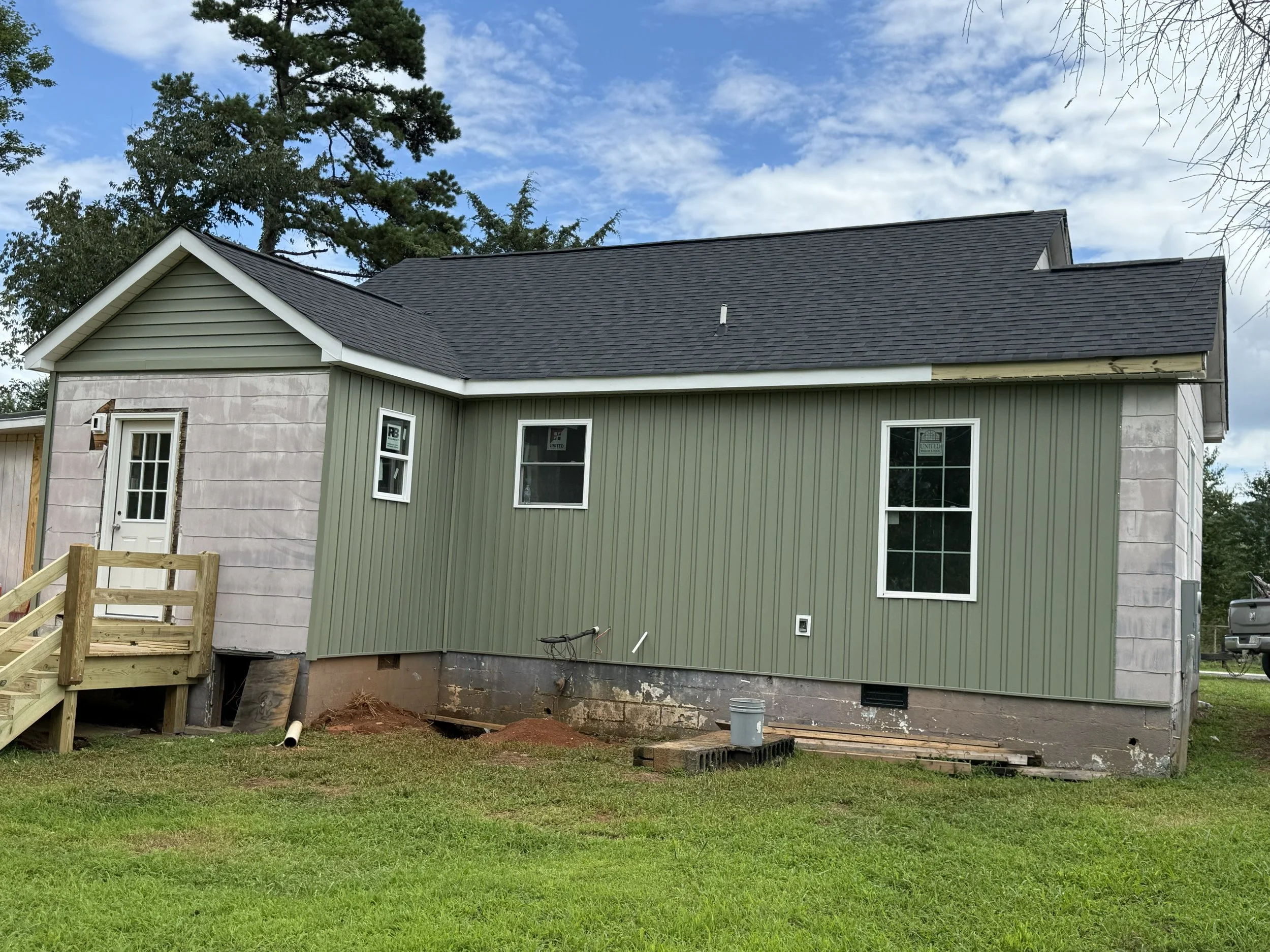 Under construction house with partially finished green siding, staircase, and construction materials in the yard beneath a partly cloudy sky.