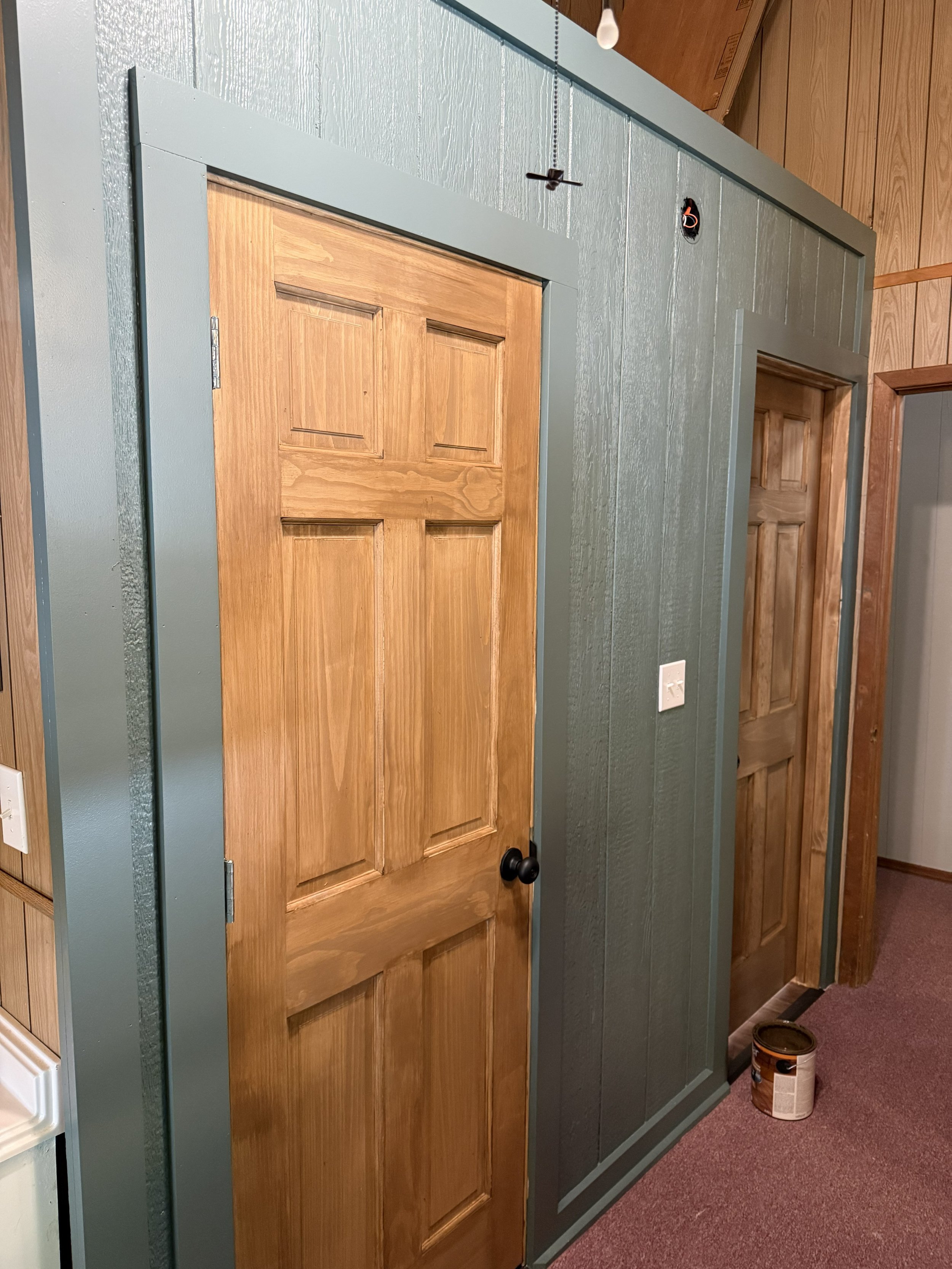 A wooden door with a dark doorknob in the center of a painted wall. The door is partially open, revealing another door behind it. The wall is painted in a muted blue-green color, and there is a white electrical outlet near the bottom right of the ima