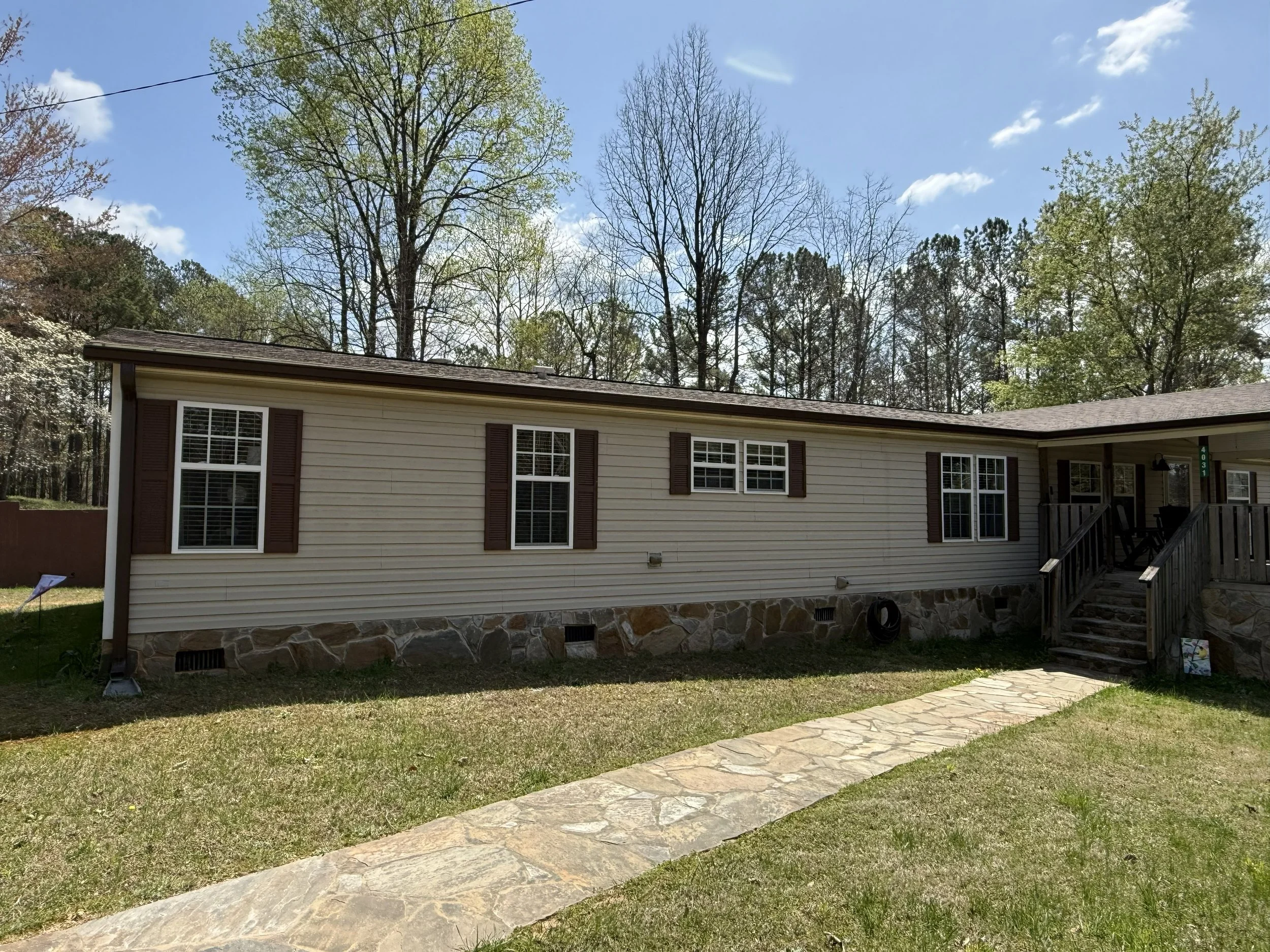 A beige house with brown shutters and a stone foundation, surrounded by a grassy yard and trees in the background. The sky is blue with some clouds.