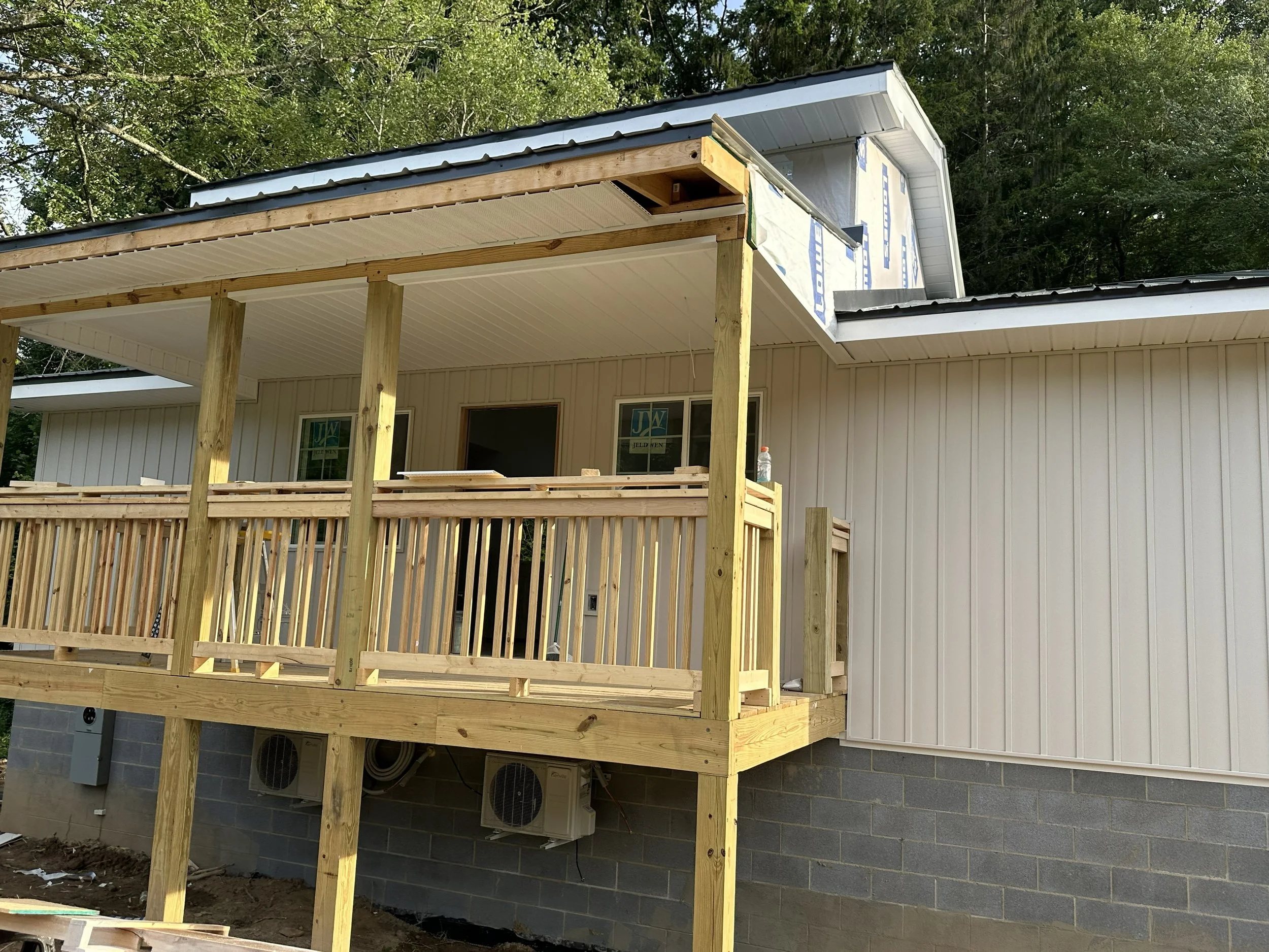 Construction site of a house showing an elevated deck or balcony under construction, with wooden support beams and railing, attached to a building with white siding and a metal roof, surrounded by trees.