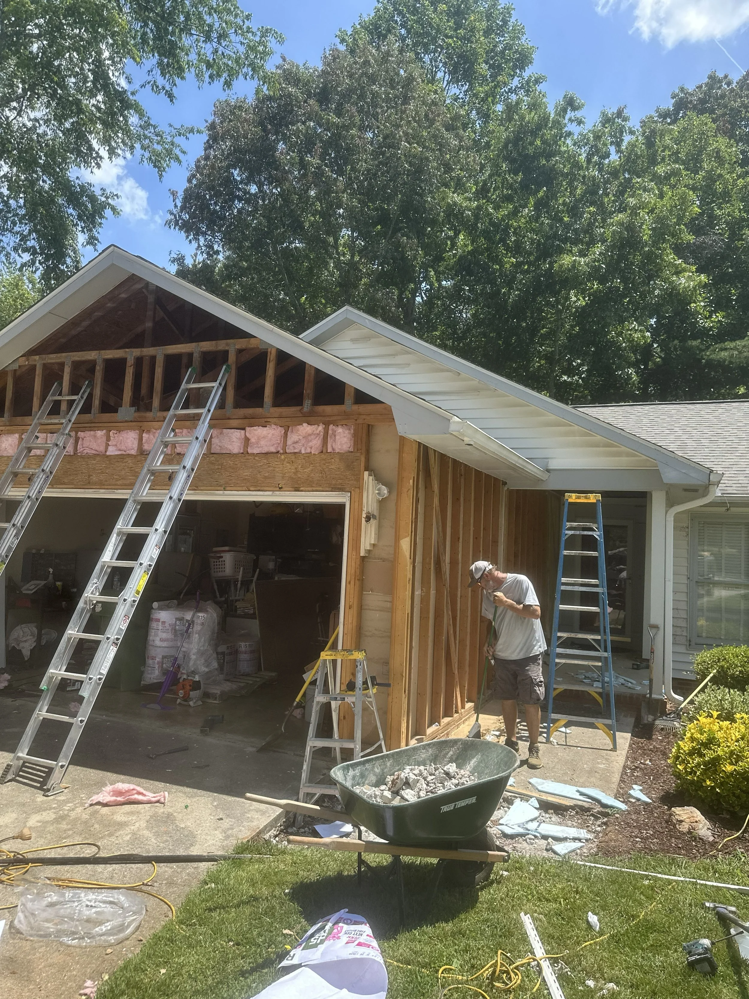 Construction workers build a house extension with a sloped roof, ladders, construction tools, insulation, and building materials under a blue sky with trees in the background.