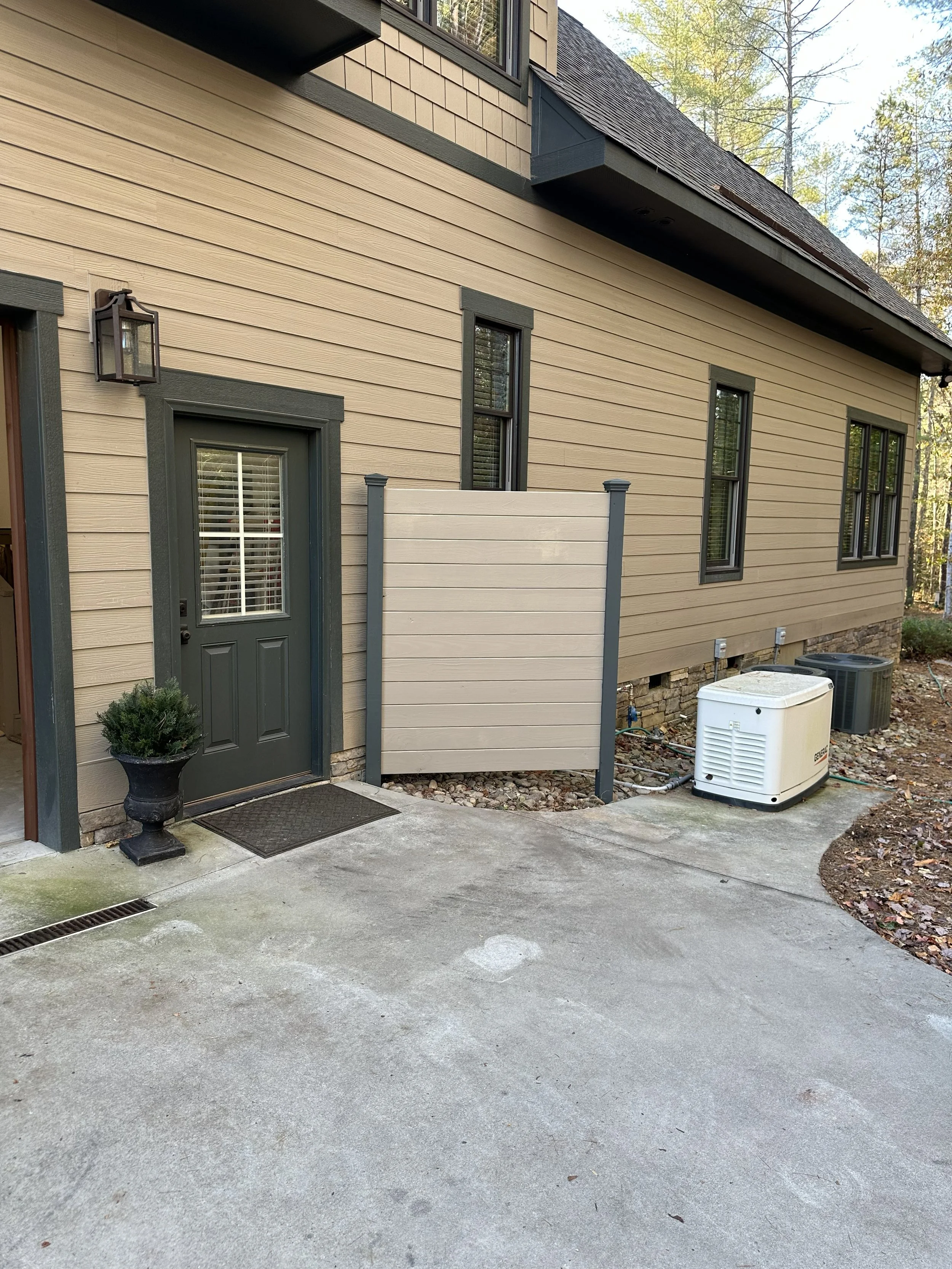 Backyard view showing a beige house with dark gray trim and four windows, a black door with blinds, a small potted plant, and outside HVAC units on a concrete surface. There are trees in the background.