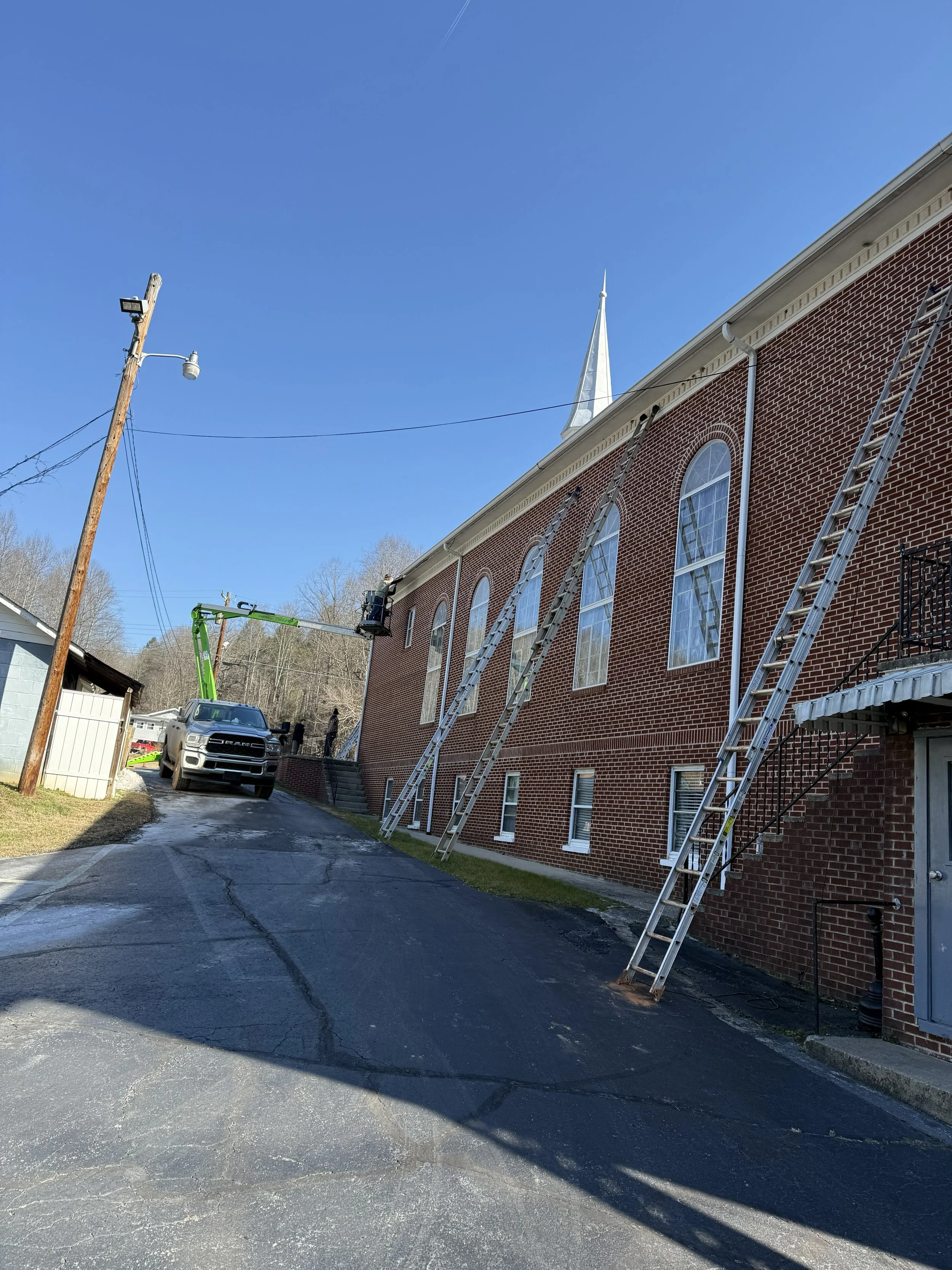 Ladders leaning against a brick church building with large arched windows, a utility pole with a street light, workers near a lifted bucket, and a truck on a paved driveway under a clear blue sky.