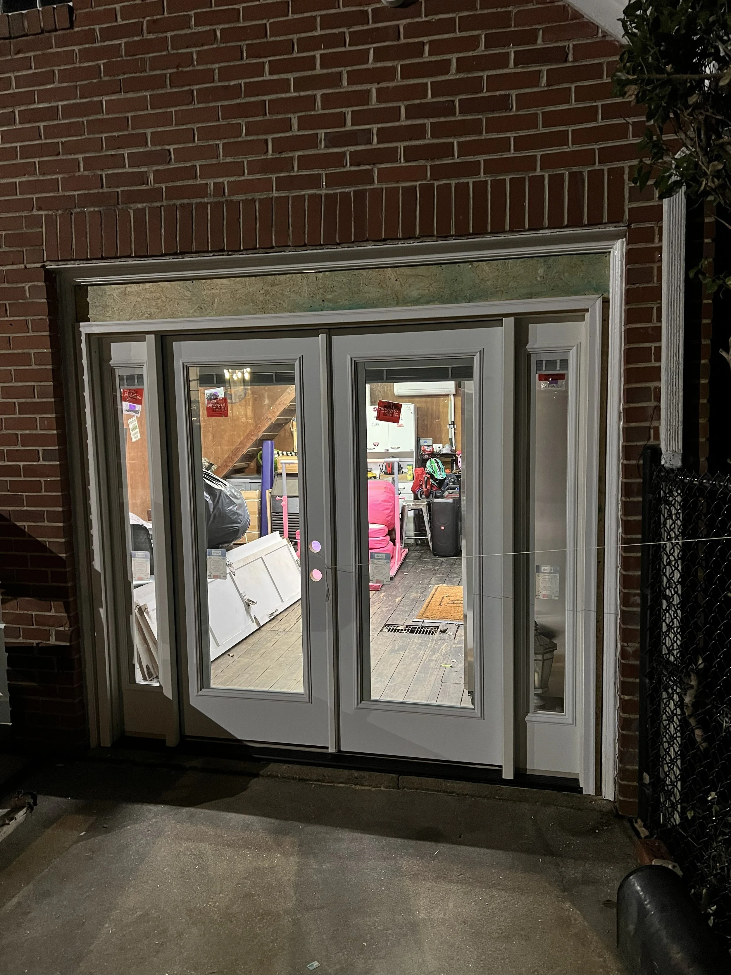 Night view of a glass door entrance to a building, with the interior visible, showing various items including furniture, bags, and equipment.