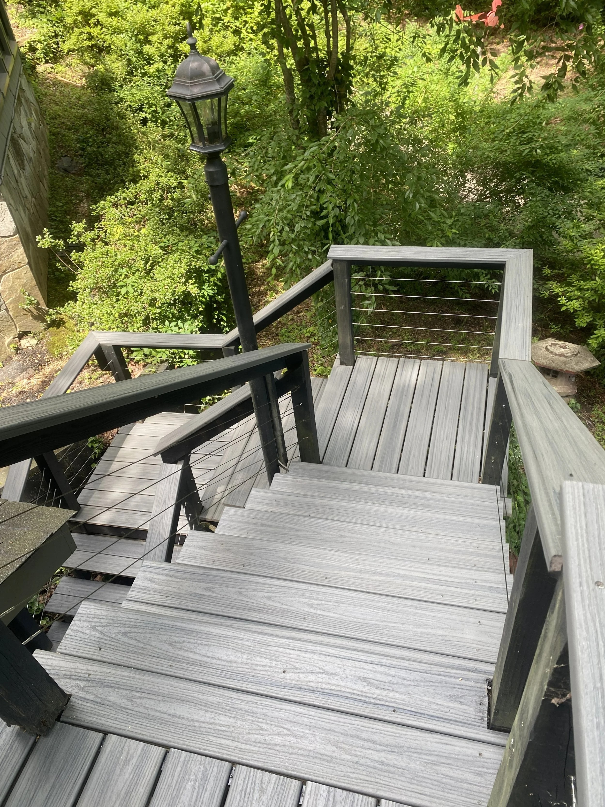 Wooden stairs leading down to a garden area with green bushes and trees, a black lamp post near the stairs, and a small stone structure or bird bath at the bottom right.