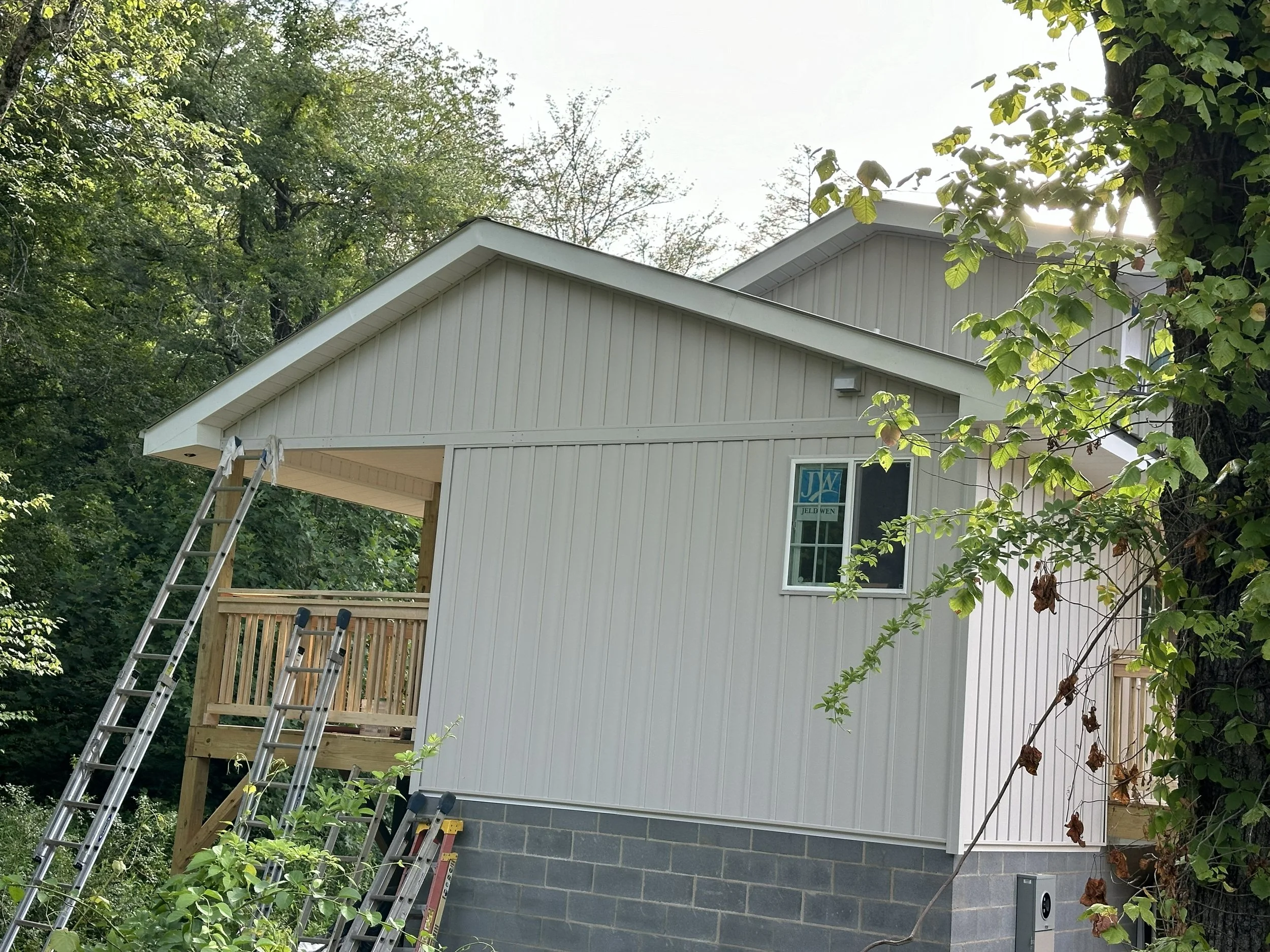 Back of a house under construction, with a staircase and a partially built porch, surrounded by trees and greenery.