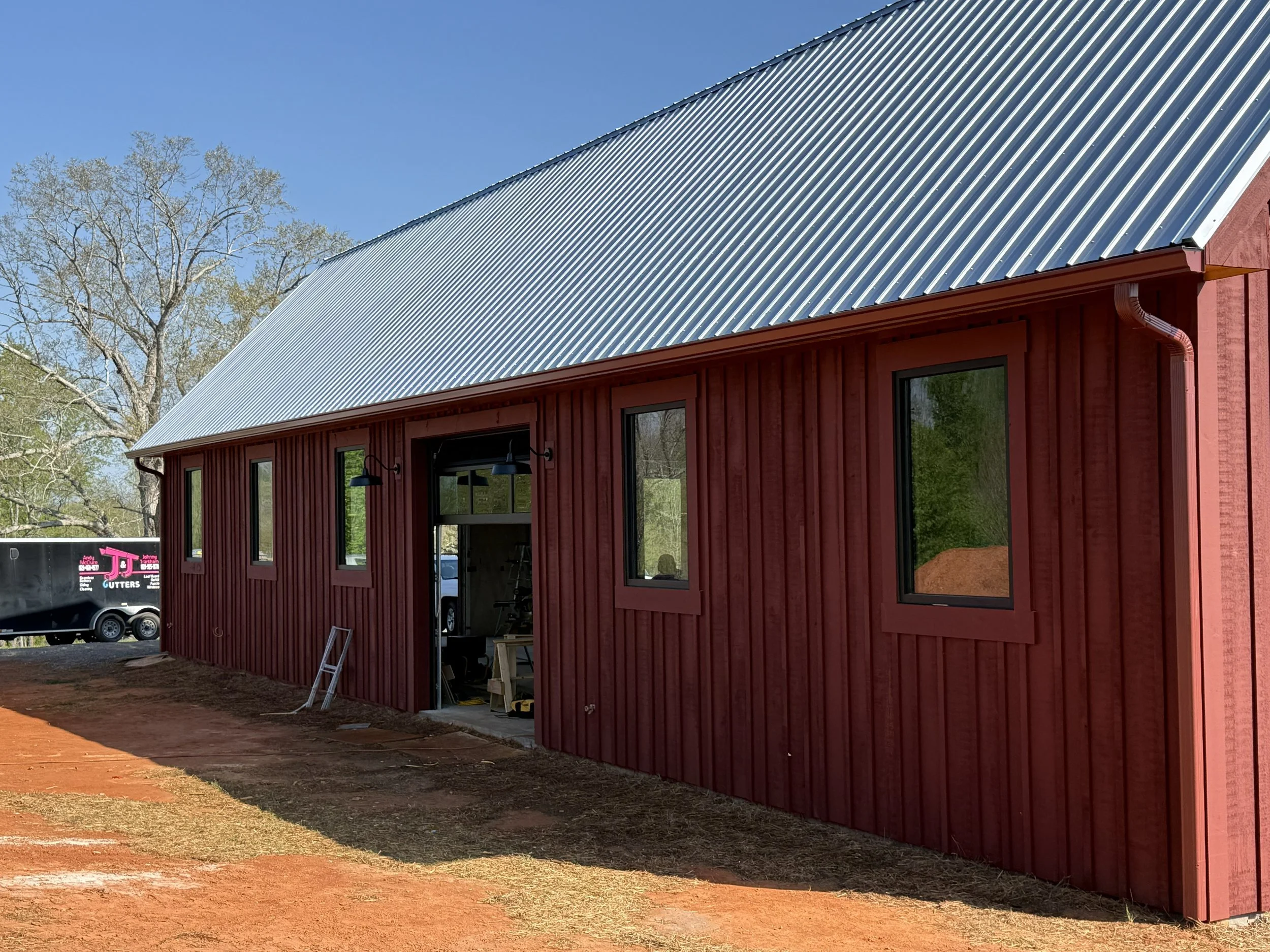 Red barn under construction with metal roof, large windows, and a dirt yard.
