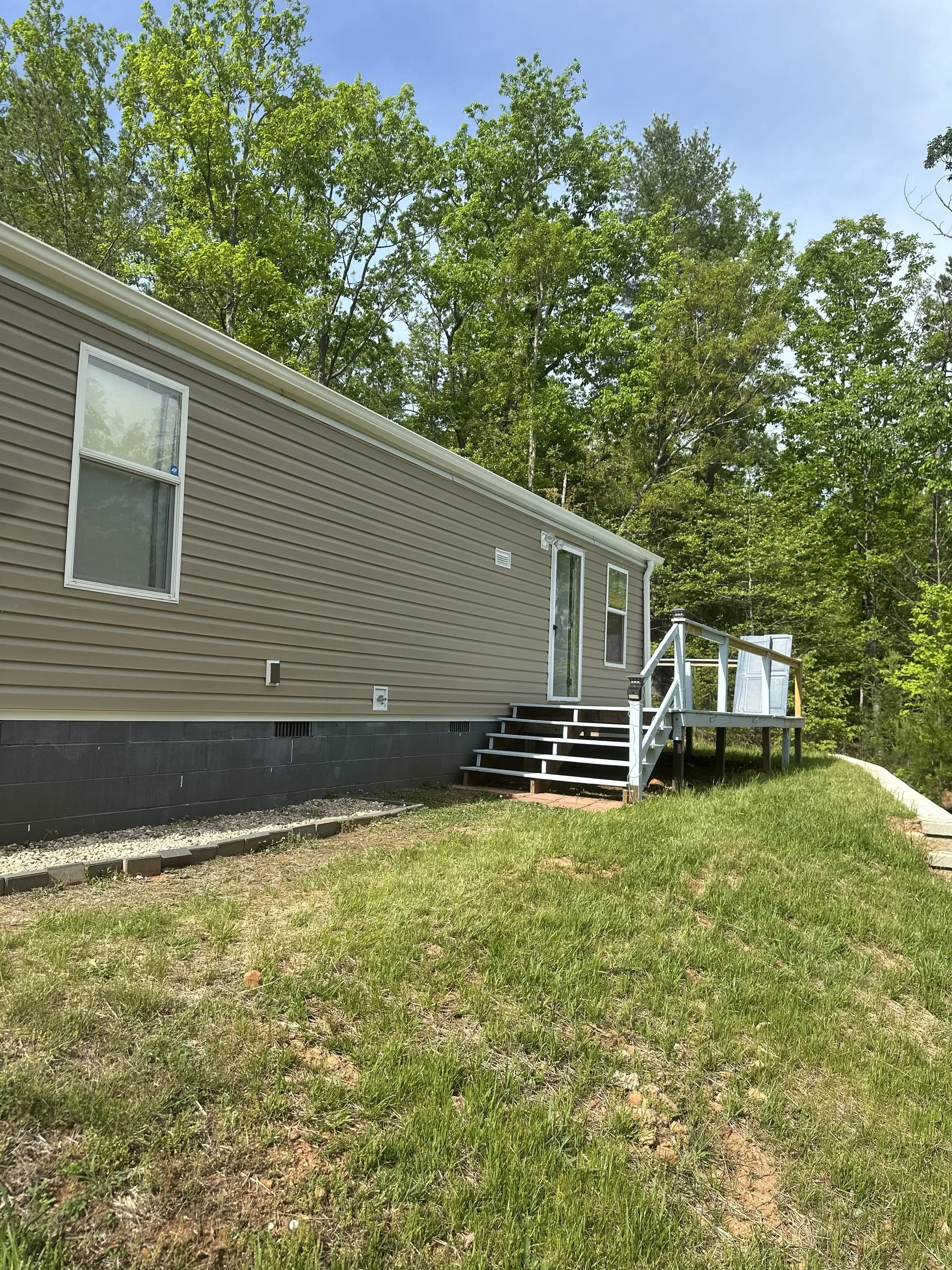 A beige manufactured house with white trim, two windows, and a small wooden staircase leading to a deck, surrounded by green grass and tall trees under a clear blue sky.