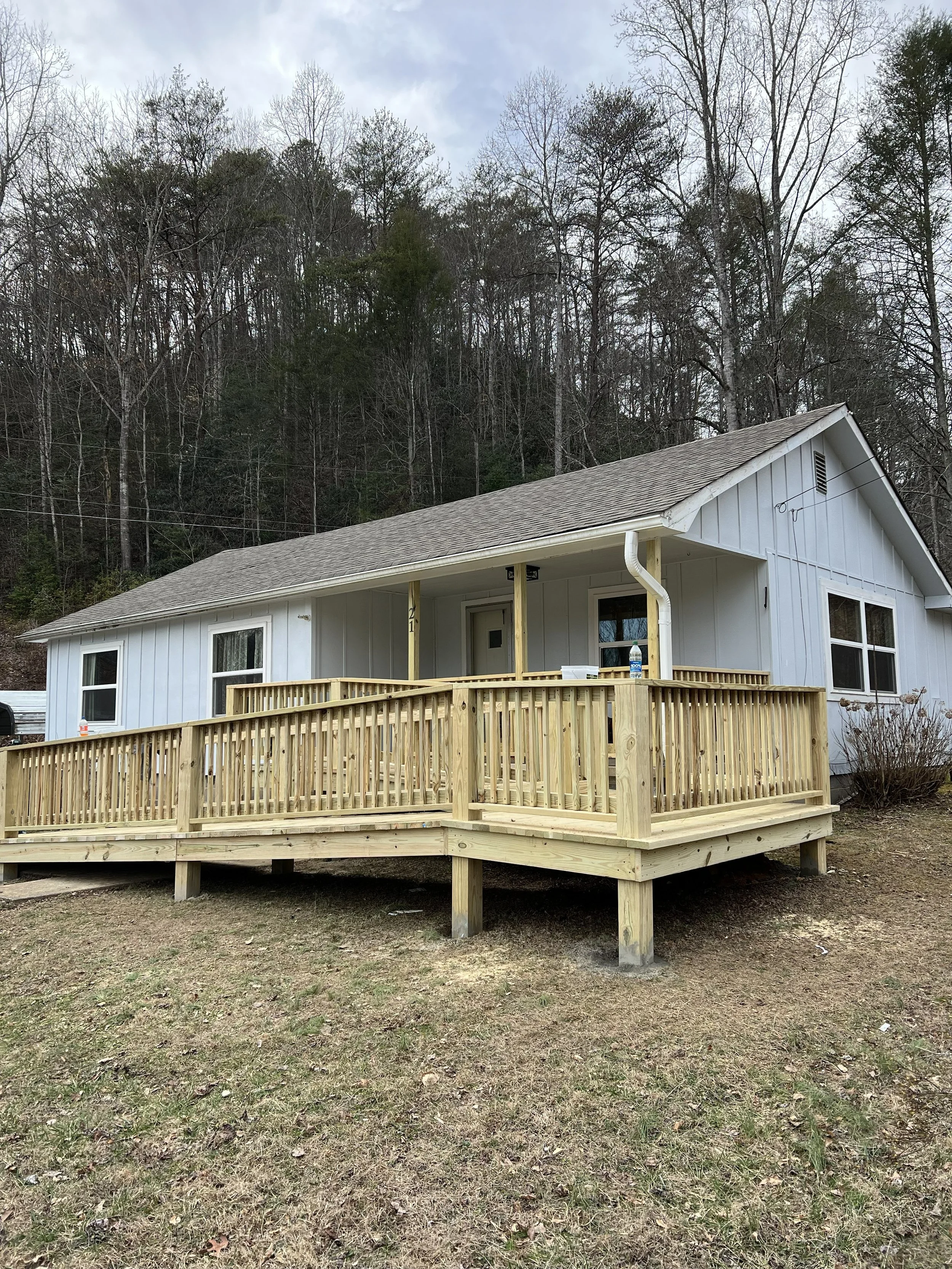 A house with white siding and a gray roof, featuring a deck under construction with unfinished wooden railings, situated in a yard with sparse grass and leafless trees in the background.