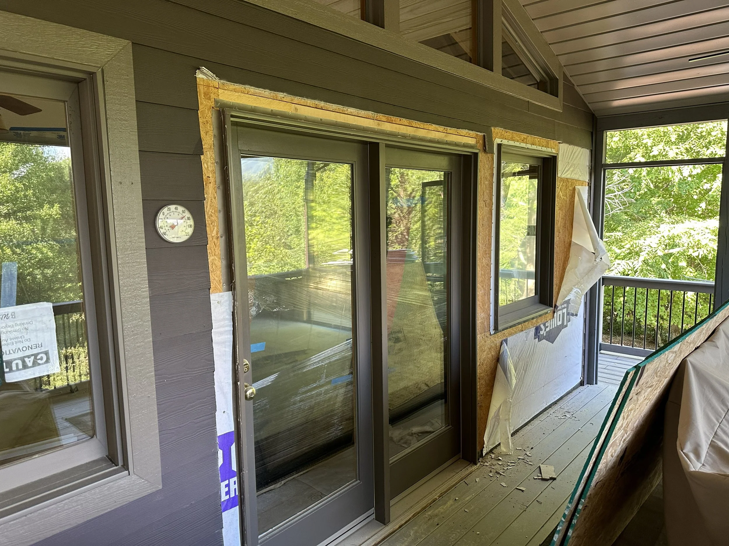 Image of a construction area with unfinished exterior wall, sliding glass doors, and windows, surrounded by trees visible through the glass.