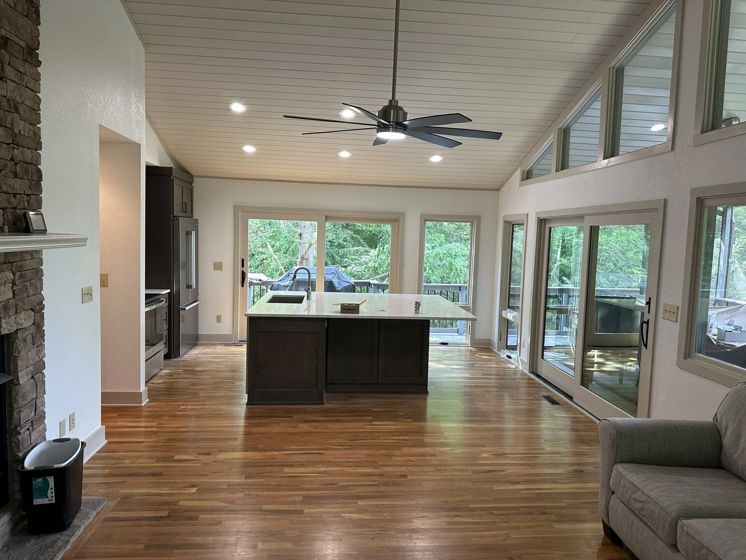 Open-concept kitchen and living room with wood floors, white walls, large windows, sliding glass door, and a ceiling fan.
