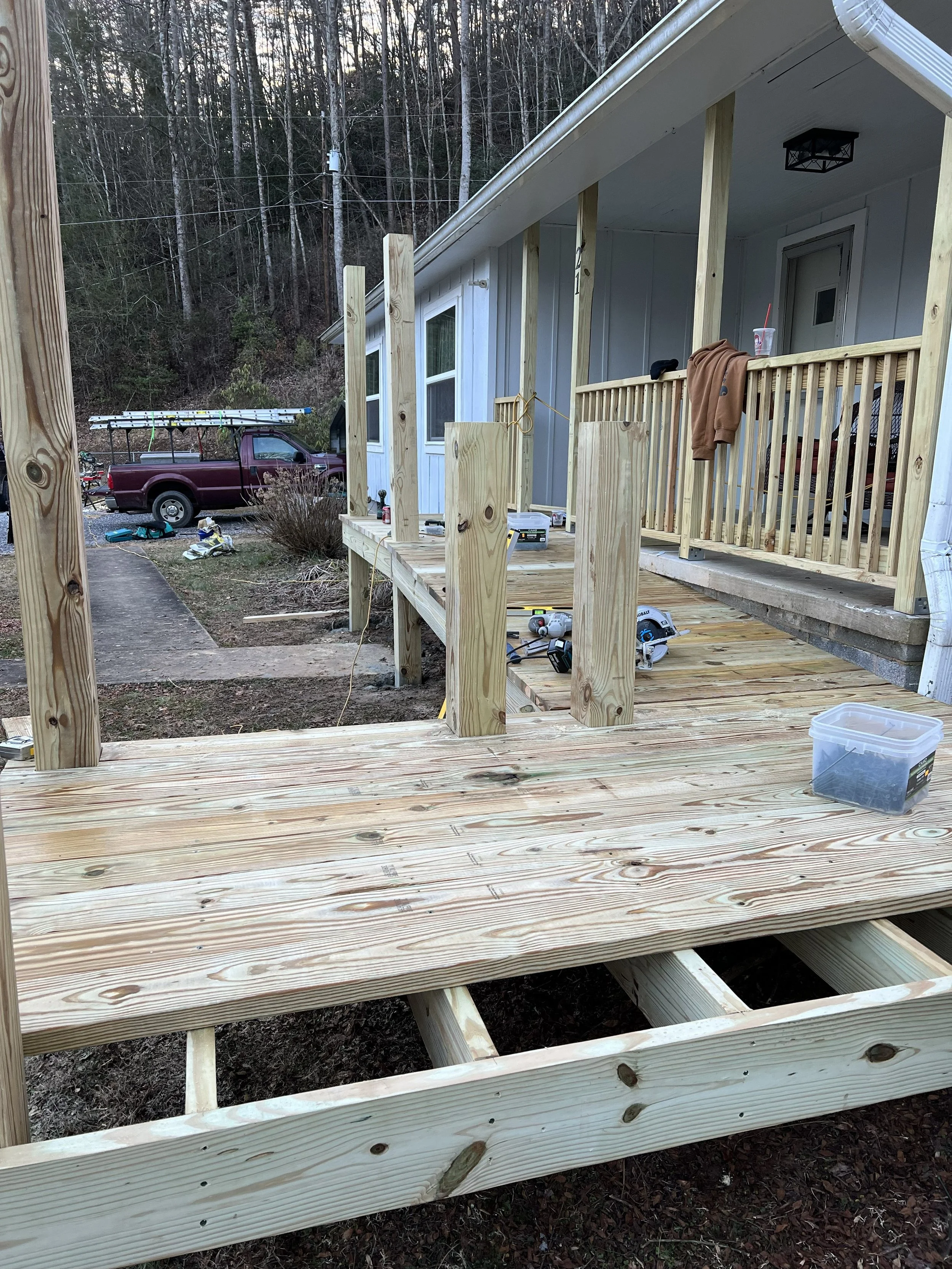 Wooden porch railing being built on a house with a white exterior, with a truck and tools in the background, on a wooded hillside.