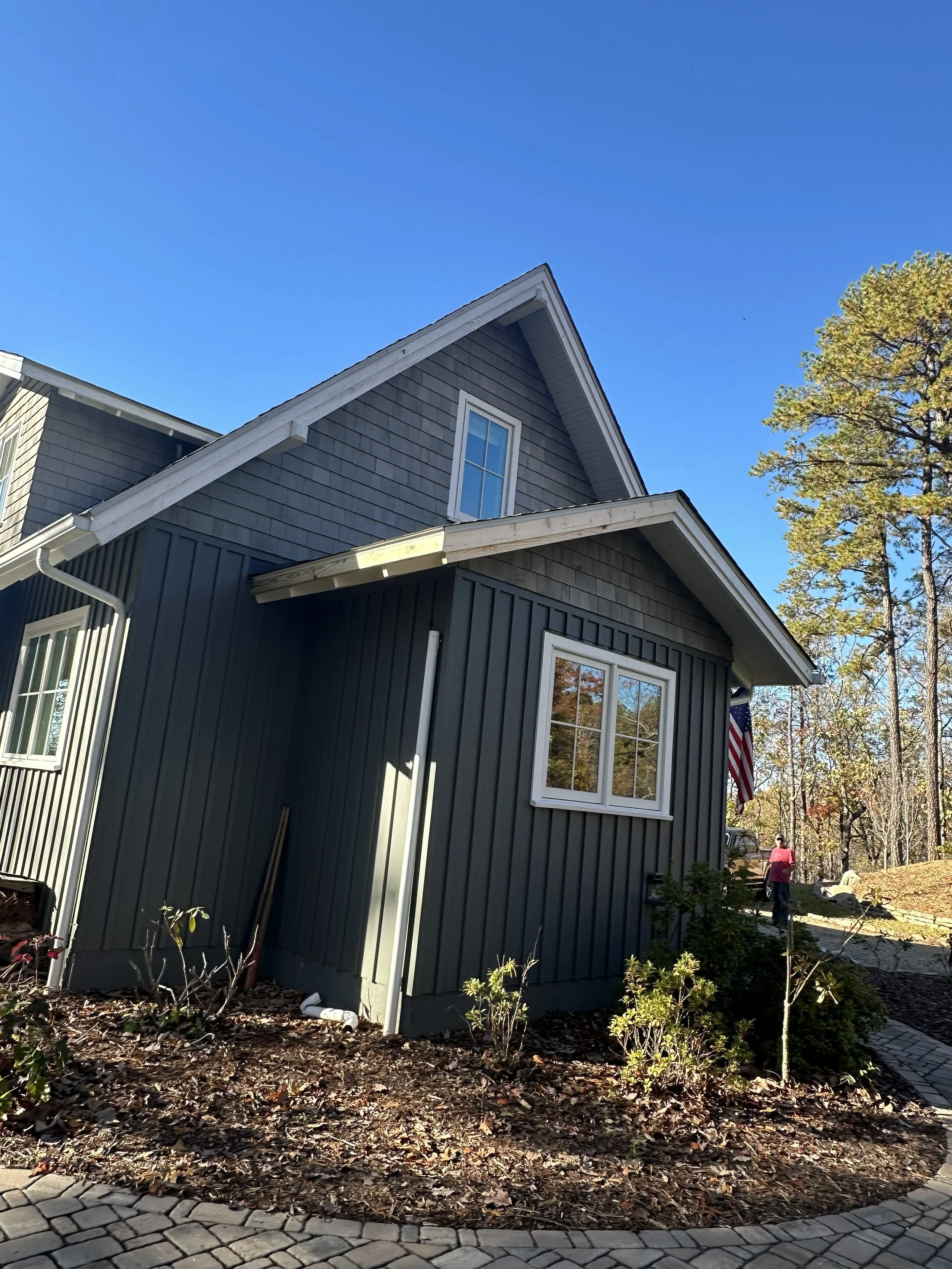 A two-story house with dark gray siding, white trim, and a gabled roof under a clear blue sky. There are windows on the upper and lower floors, and a garden bed with plants and mulch in the foreground. A person is walking in the background near trees