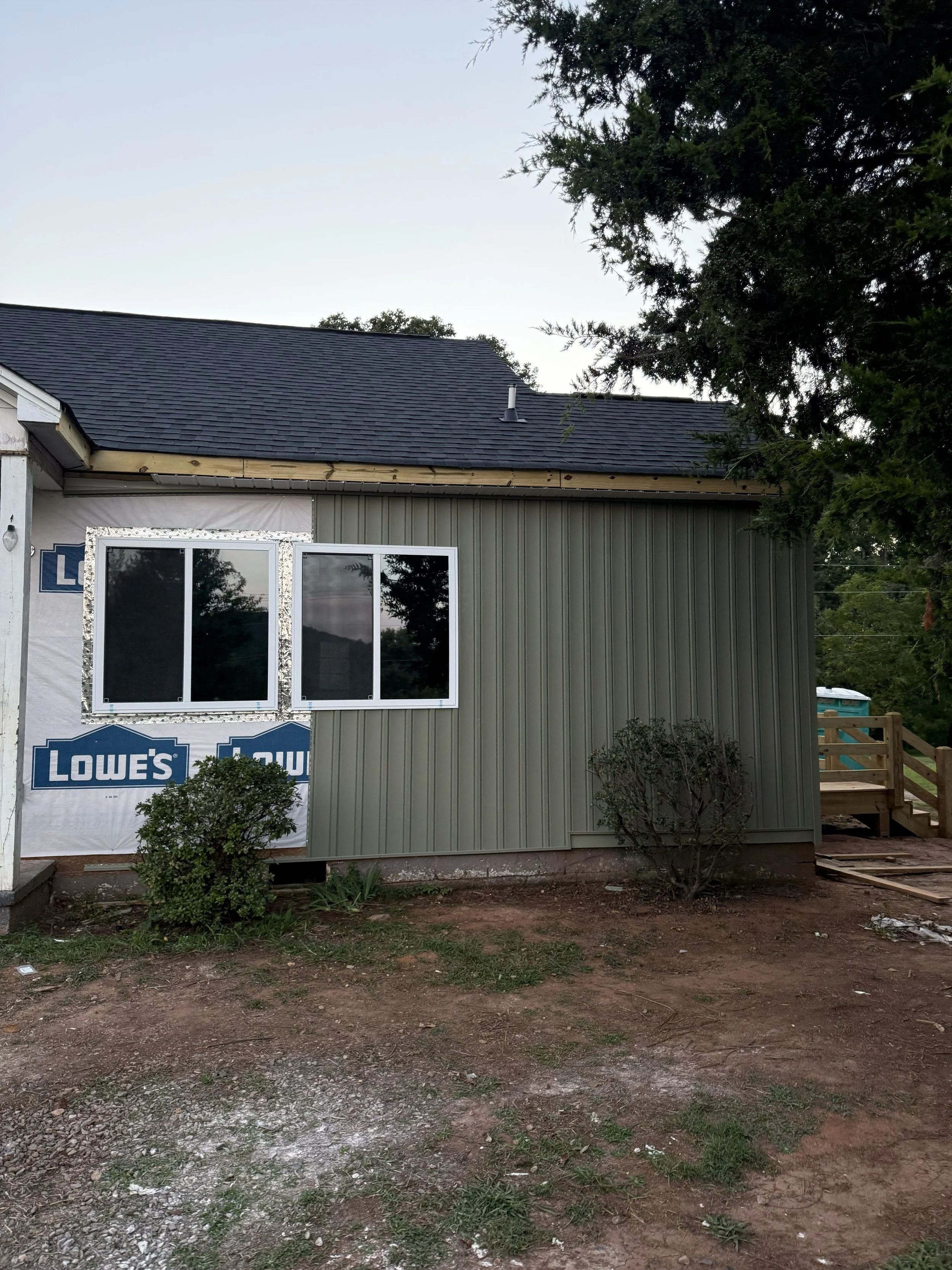 House under construction with siding installed on part of the wall, windows, and roofing completed, outdoor yard with dirt and patches of grass, trees and a clear sky in the background.