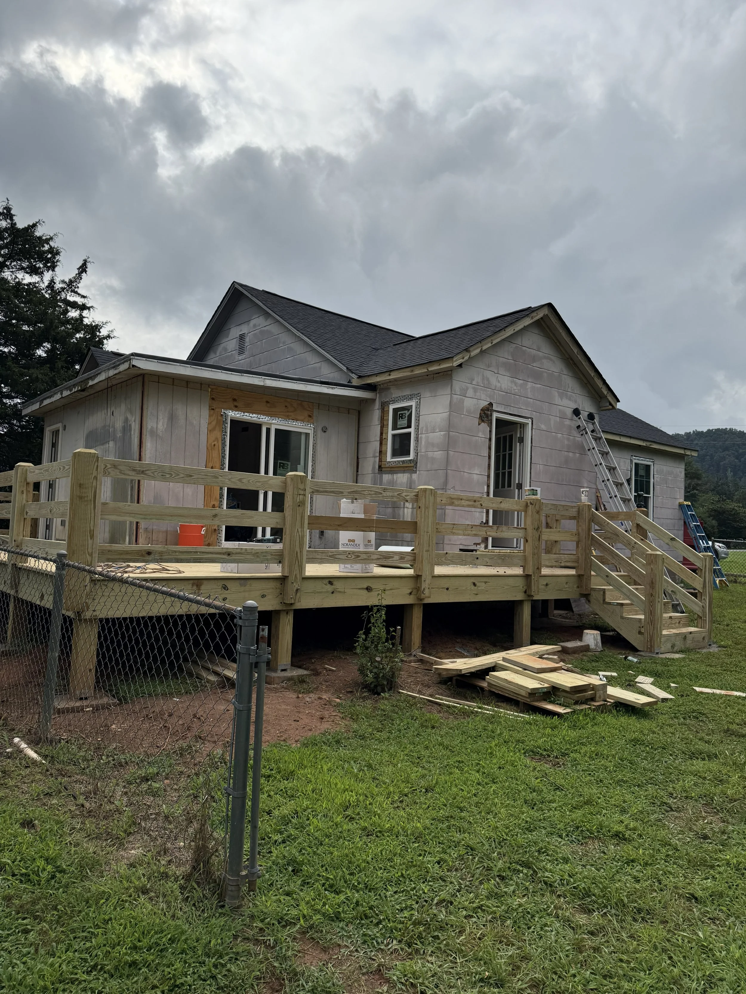 A house under construction with a wooden deck and stairs, with building materials and tools nearby, and cloudy sky overhead.