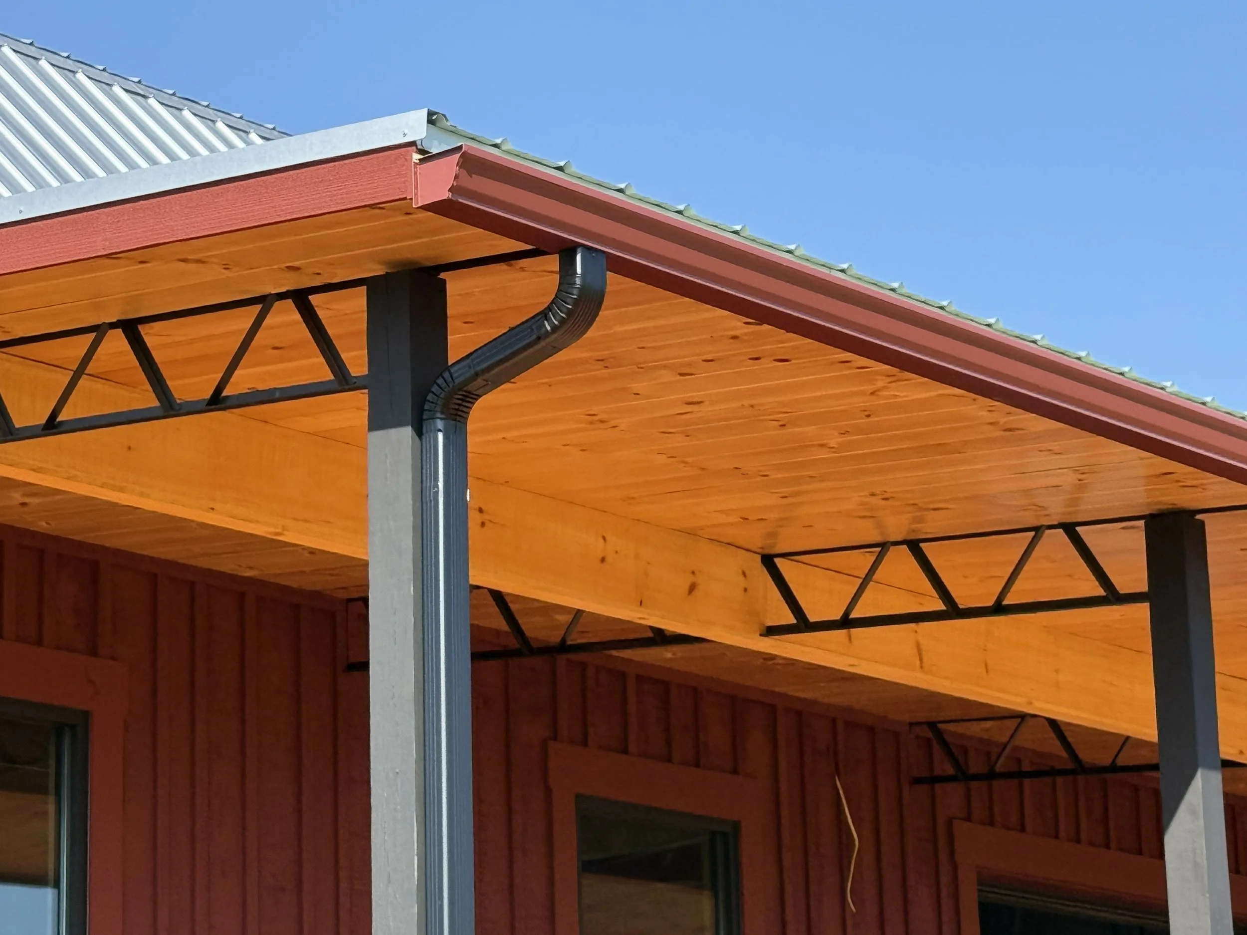 Part of a house with brown wooden exterior walls, a sloped metal roof, metal gutters, and visible black metal support beams underneath the roof. There is a clear blue sky in the background.