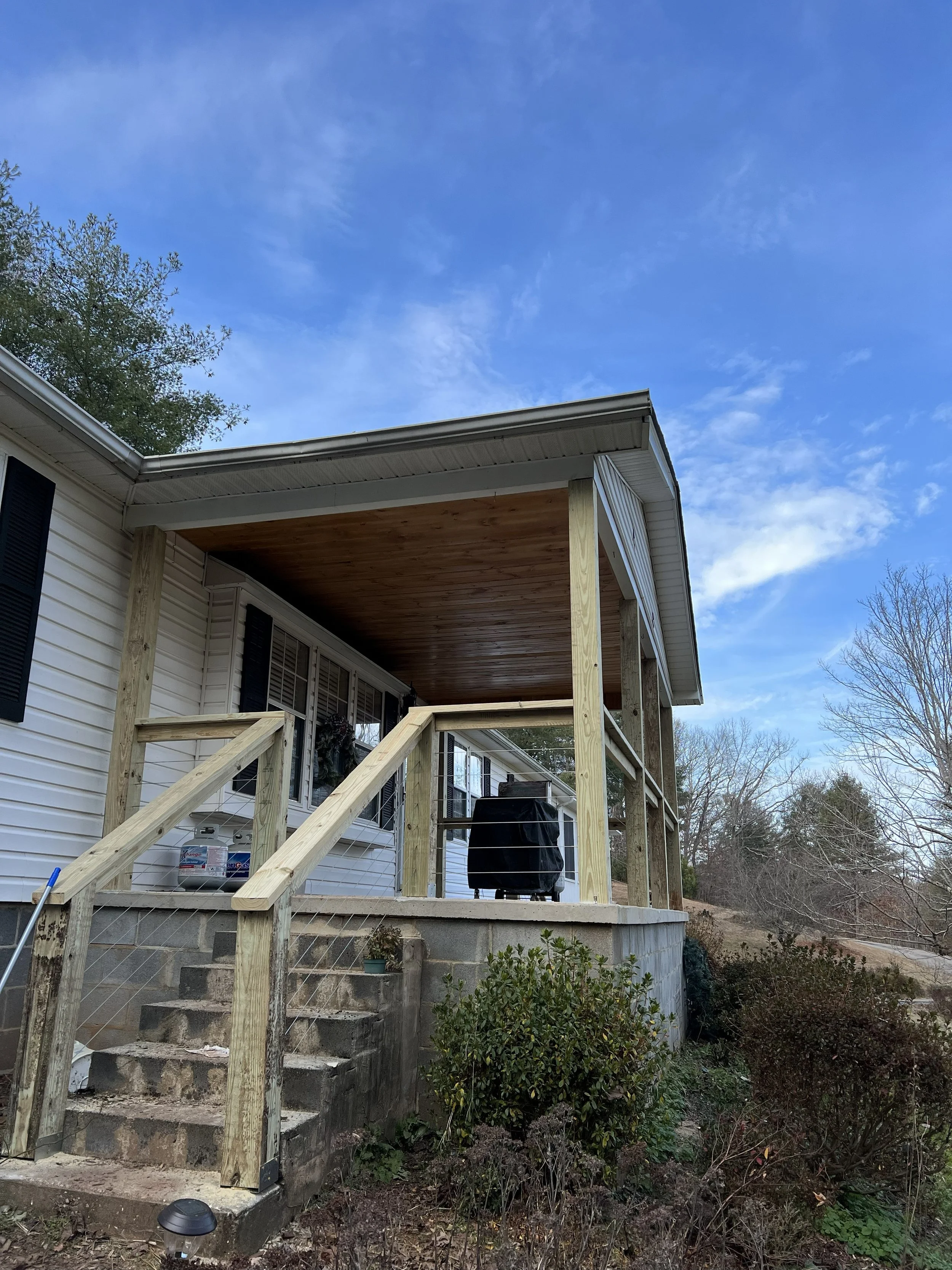 Backyard with a wooden deck under construction, with stairs and railing, attached to a house with white siding and black shutters, surrounded by trees and bushes, and a blue sky with some clouds.
