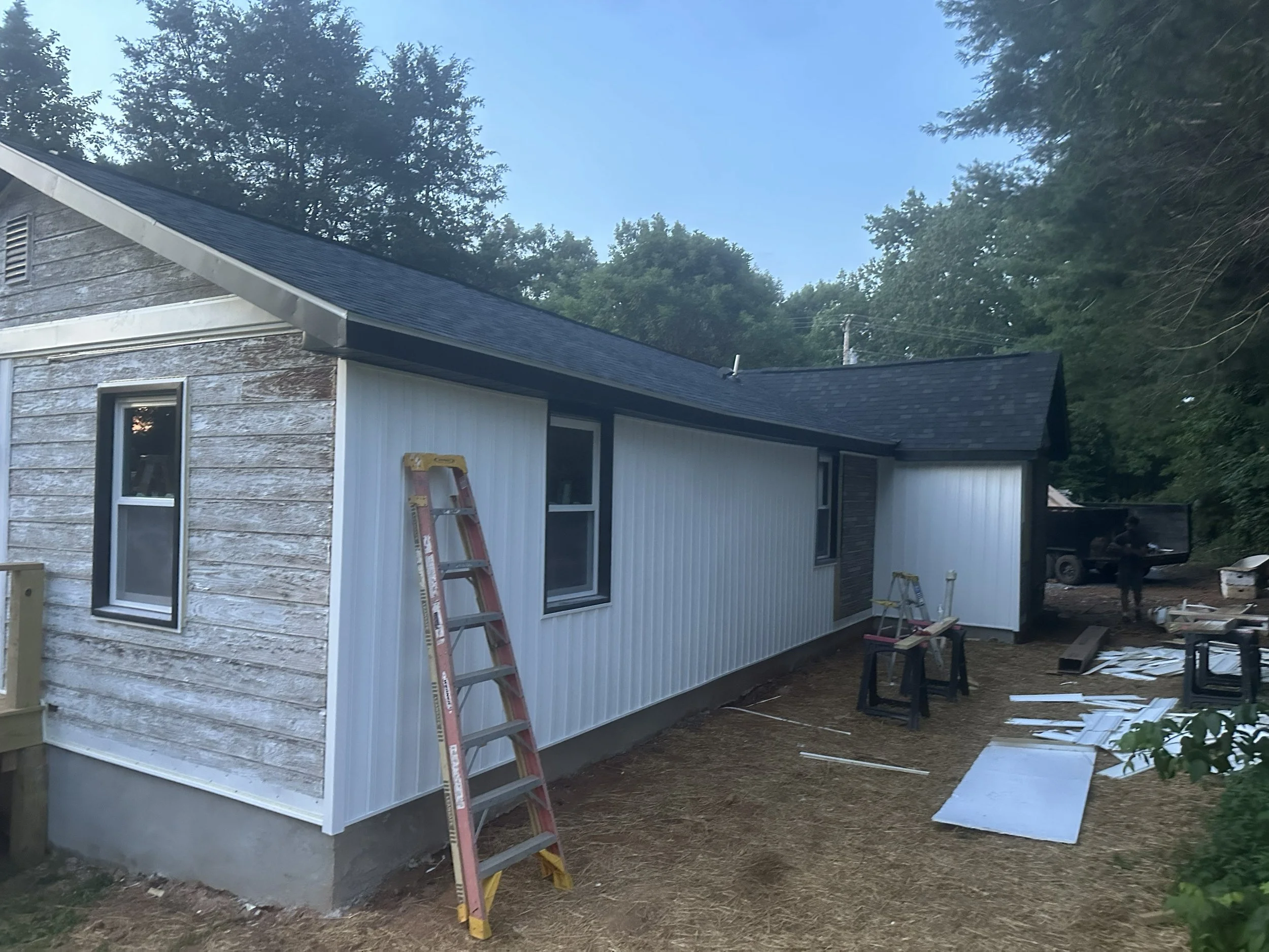 Partially constructed house with white siding, window, and ladder outside. Building materials and tools on the ground, trees in the background, and a clear sky.