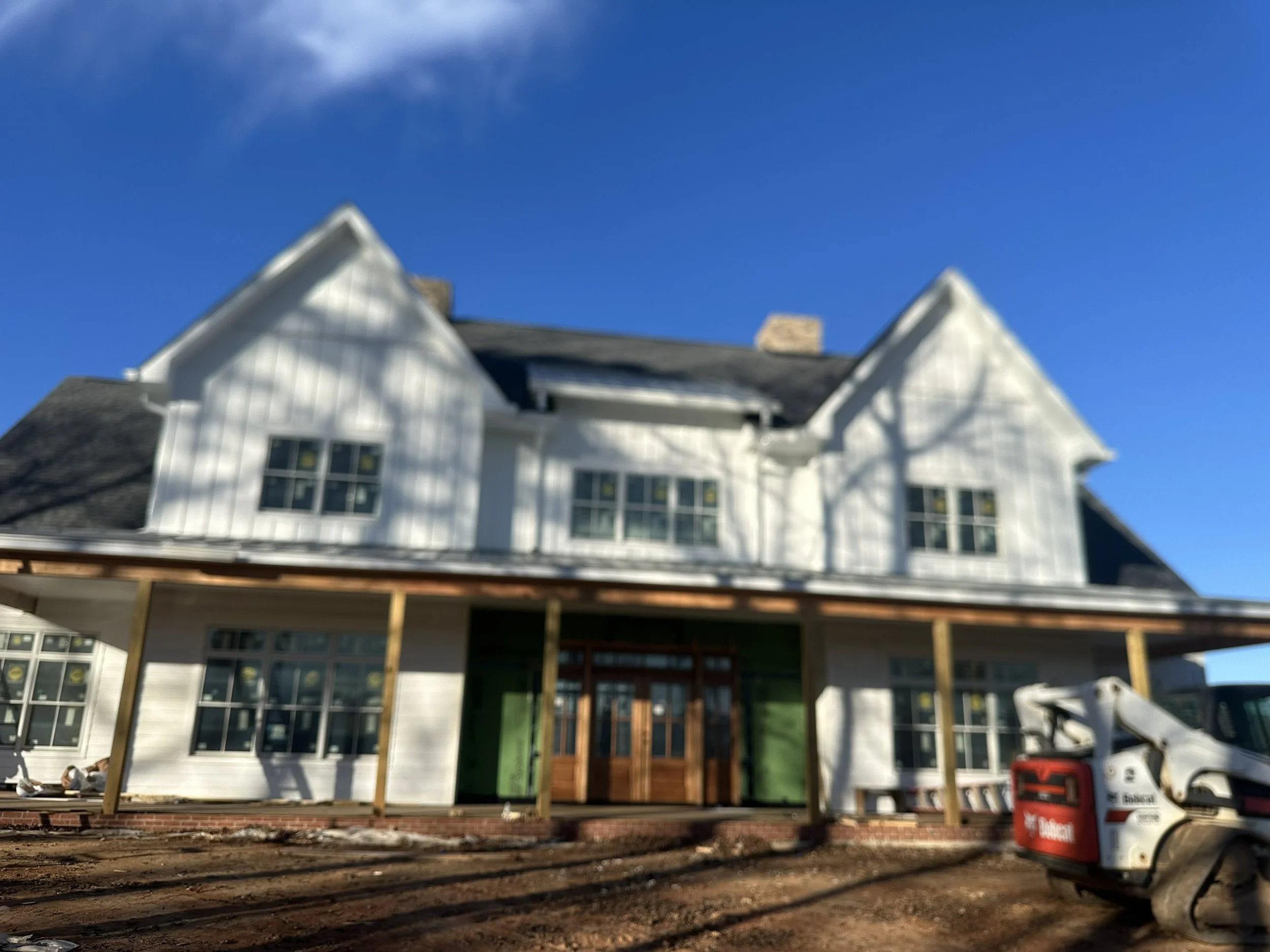 A house under construction with a partially built deck or porch, surrounded by dirt and construction materials, under a blue sky with some clouds.
