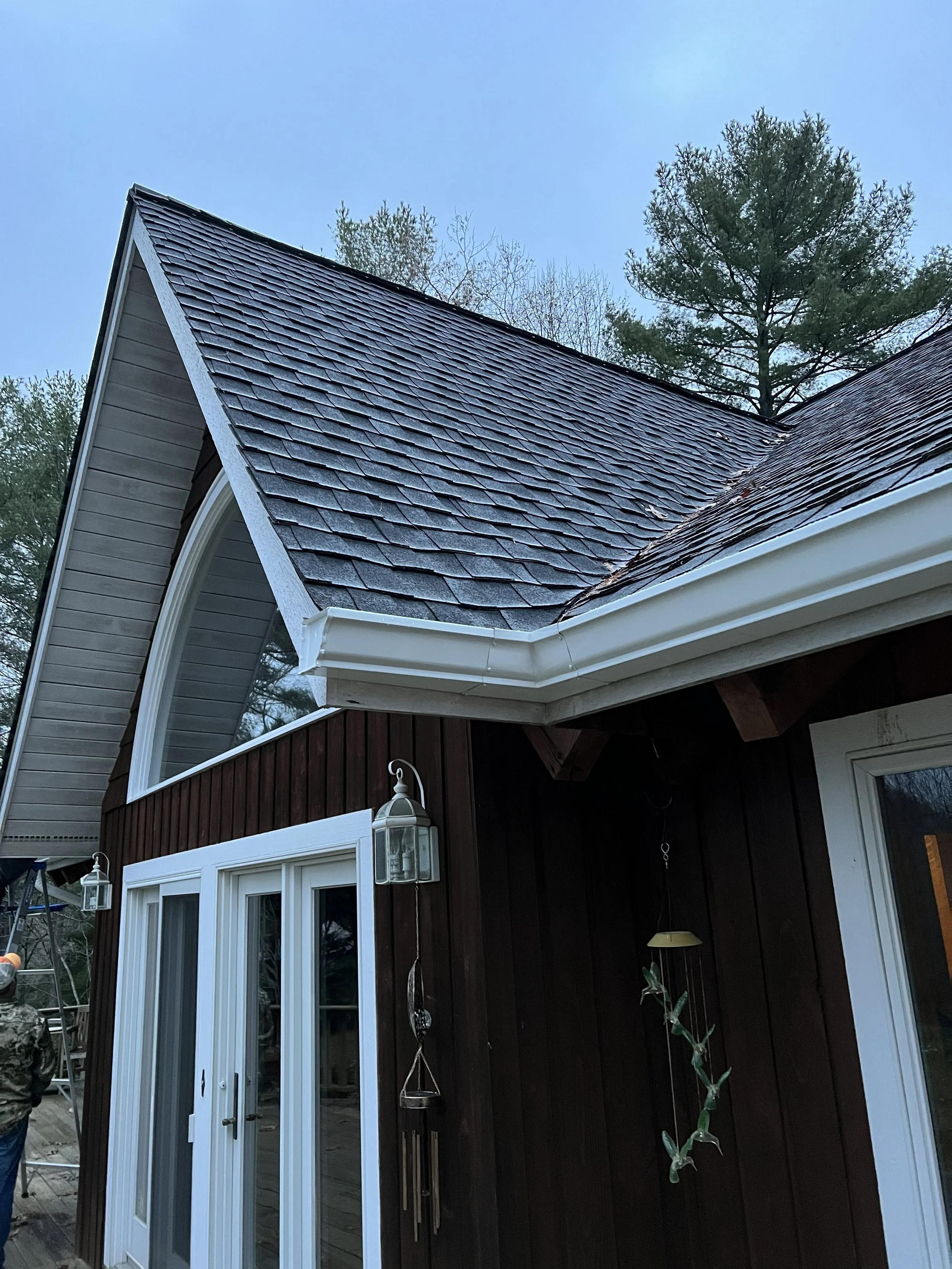 The image shows the corner of a house with a dark brown exterior wall and white trim. There are double glass doors with white frames, flanked by two outdoor lantern-style lights. The roof is steep and covered with gray shingles, with trees and a clea