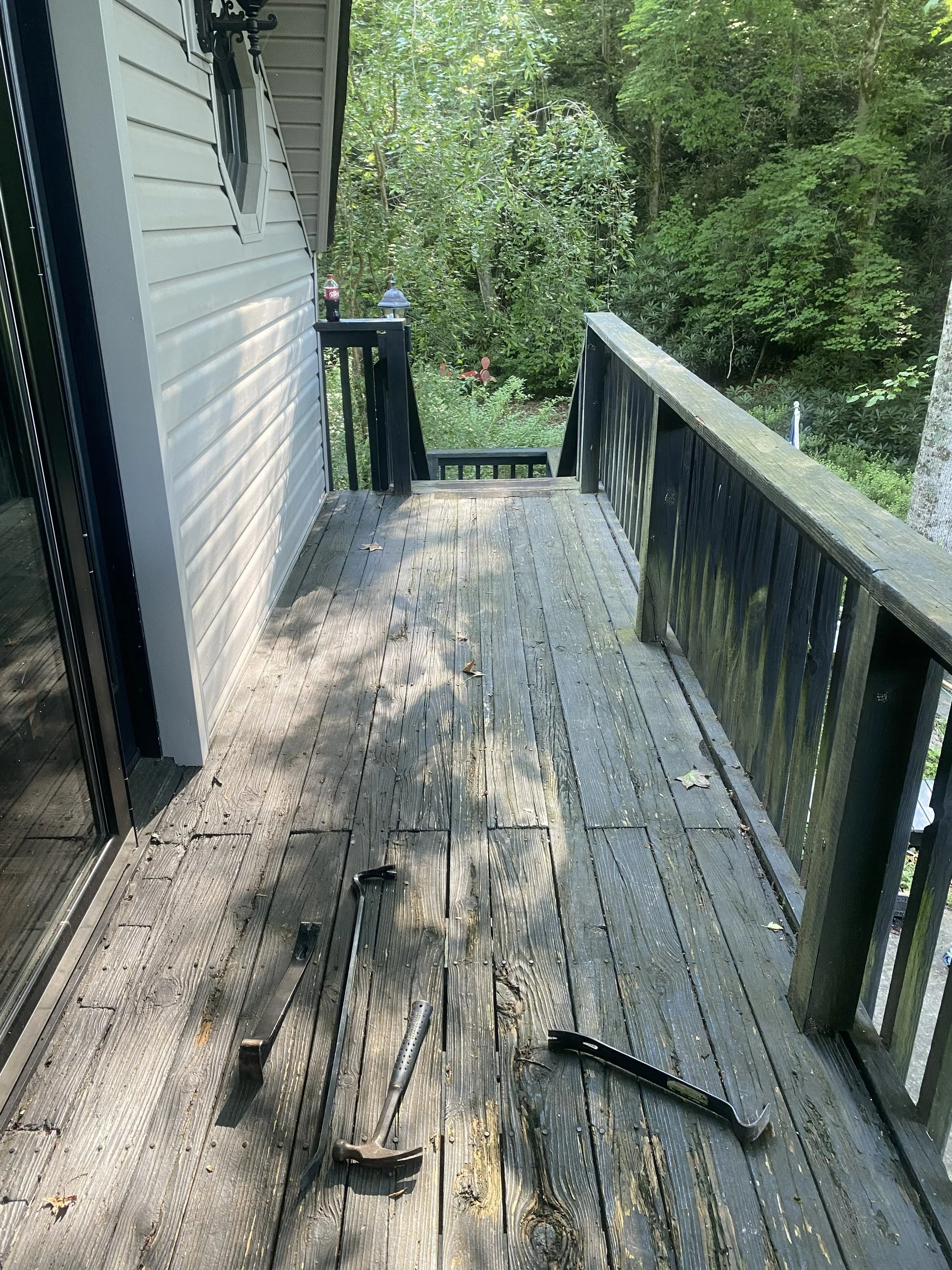 A small wooden balcony attached to a house, overlooking a lush green forest. The deck has tools and debris, including a hammer and a crowbar, indicating ongoing or recent repair work.