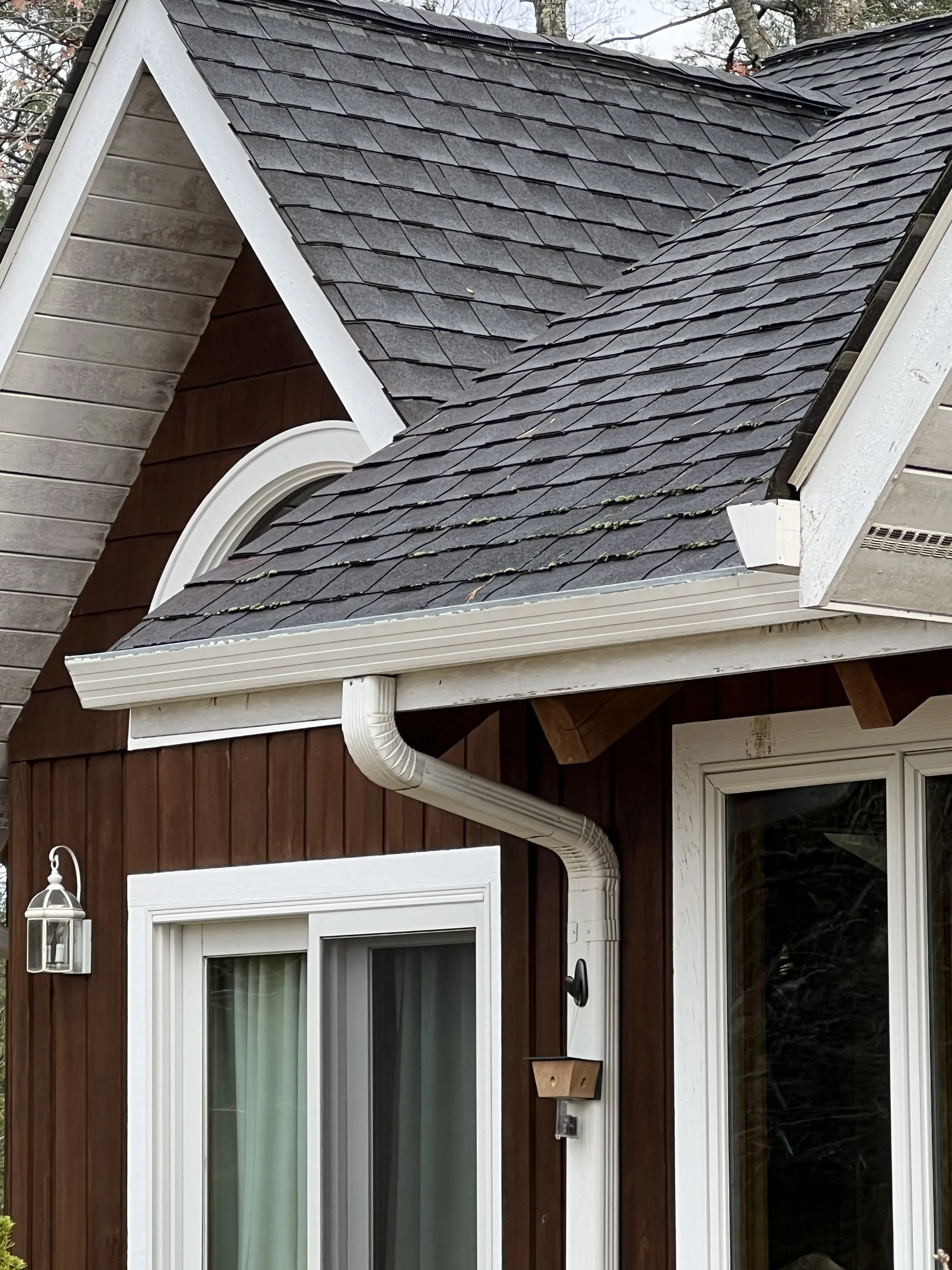 Close-up of house exterior with dark gray shingle roof, white trim, brown wooden siding, and a window with cream-colored curtains.