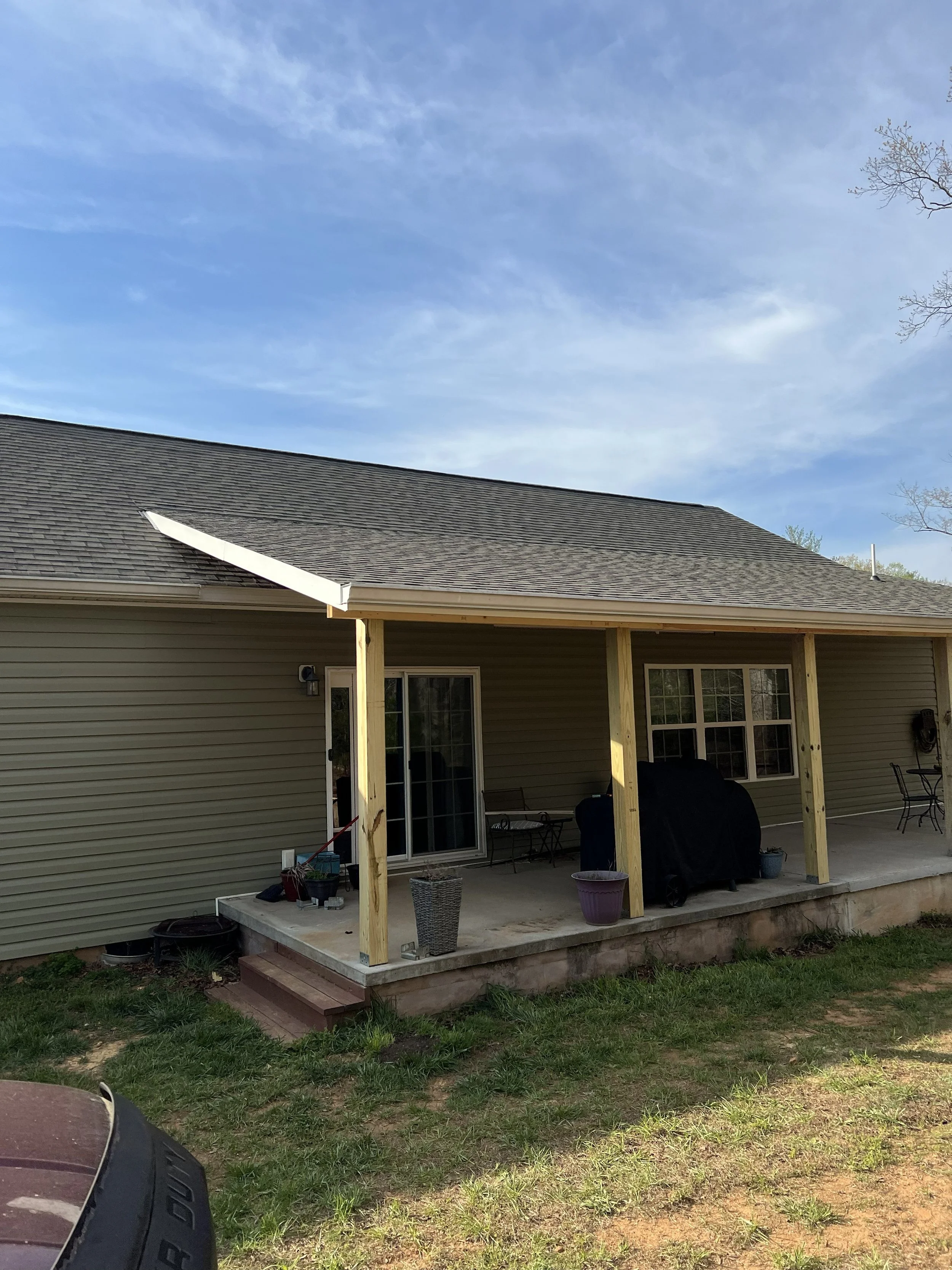 Backyard patio area with a new wooden structure under construction, with tools and plants on the concrete floor, a sliding door, and a grill covered with a black cover. The house has beige siding and a shingle roof, with trees and a blue sky in the b