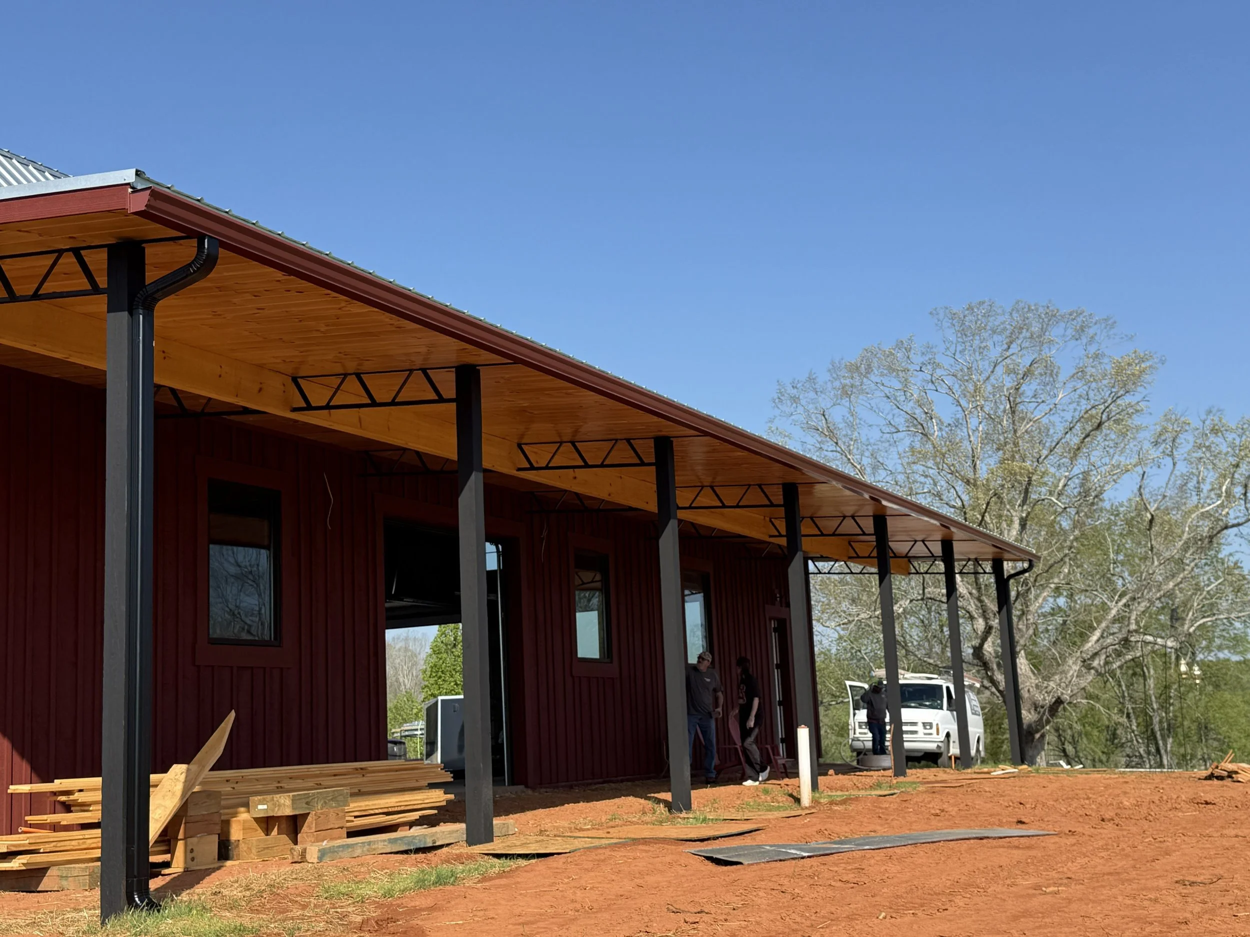 Partially constructed building with wooden and metal structure, black support beams, and a roof overhang, with a clear blue sky and trees in the background.