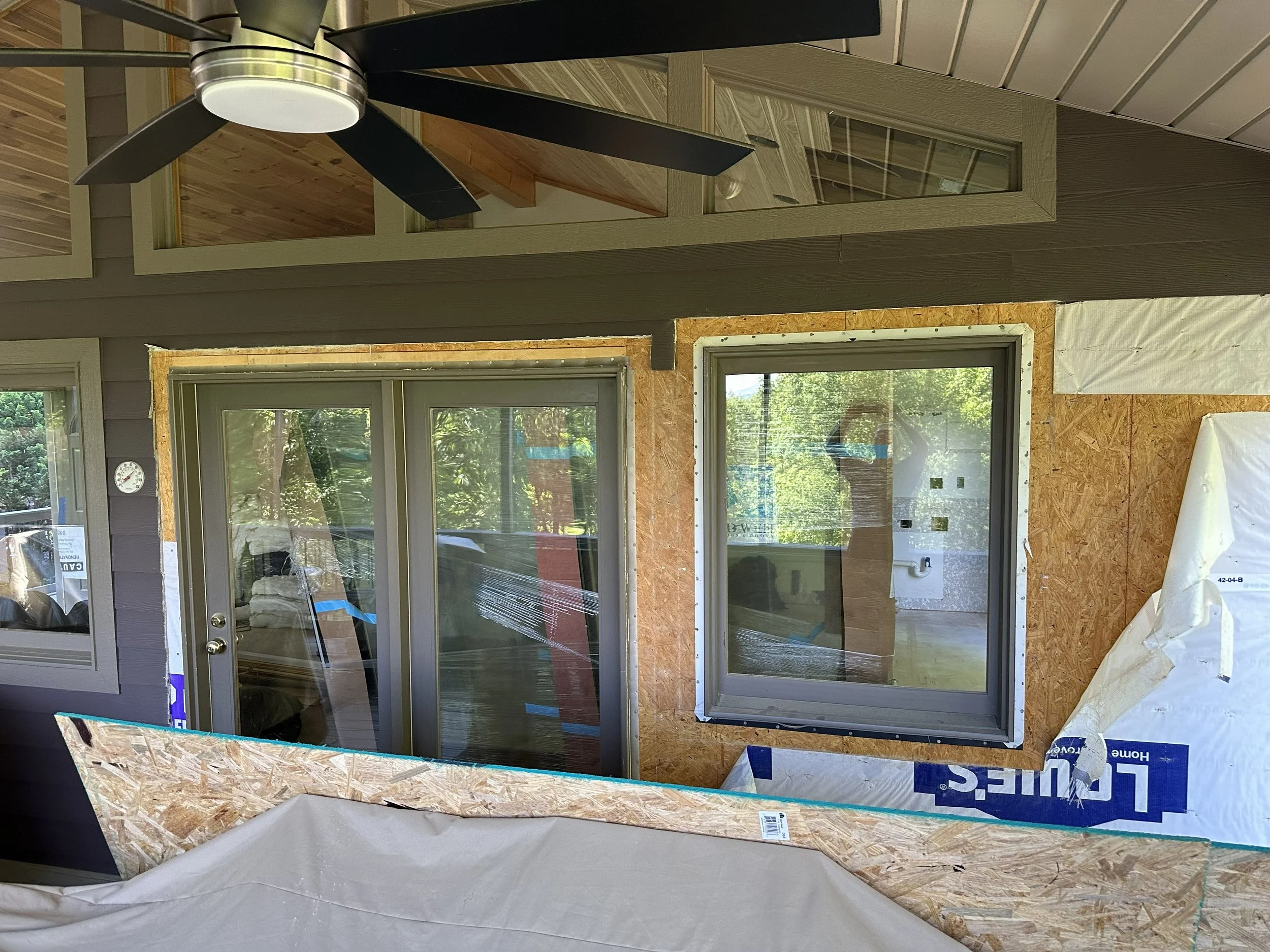 Construction site with large sliding glass doors installed on a wall made of oriented strand board (OSB) sheathing. A ceiling fan is visible on the unfinished interior ceiling. The scene shows ongoing building work with construction materials and too