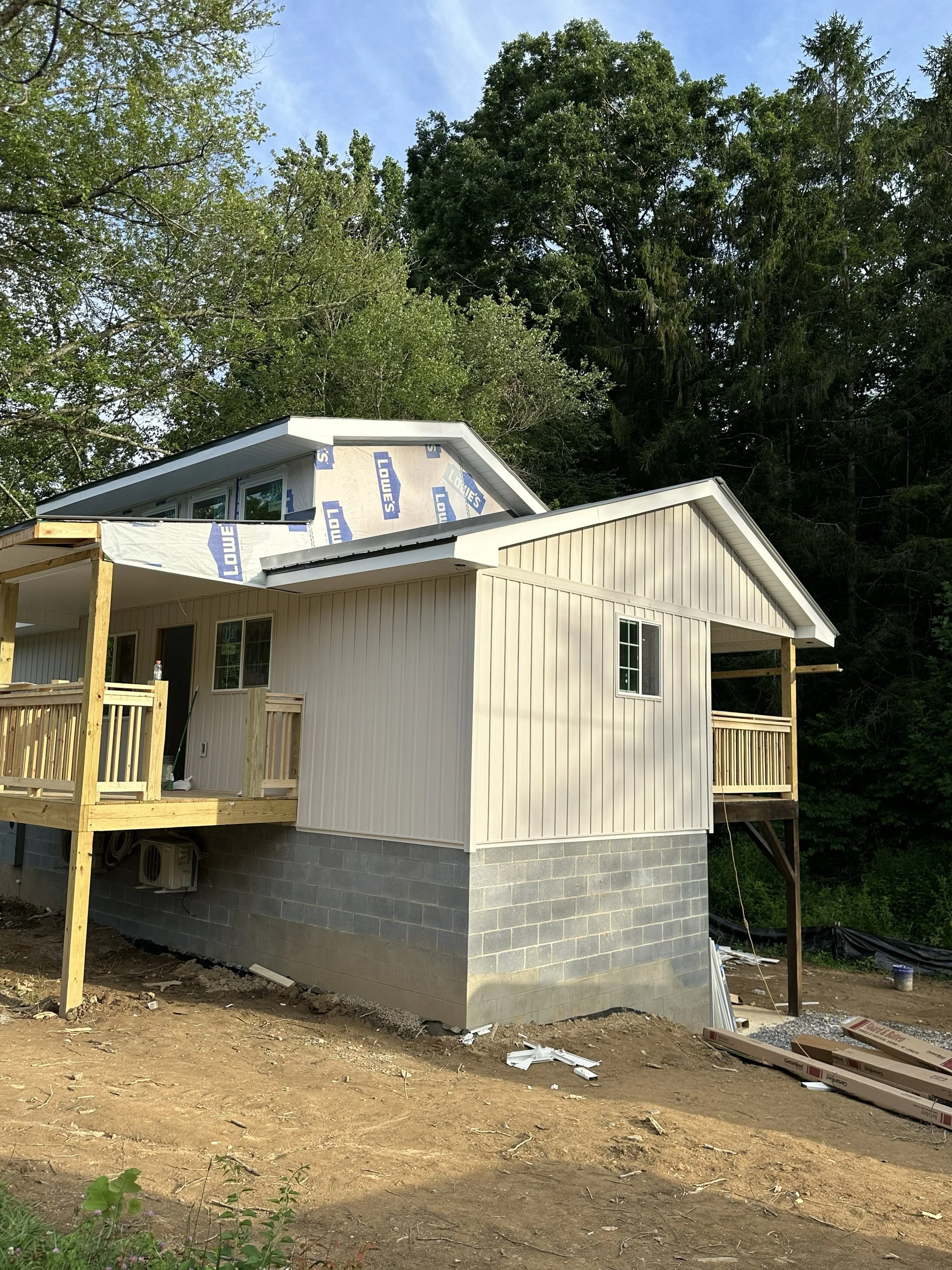 A house under construction with a partially completed exterior and a new wooden deck, surrounded by trees and a clear blue sky.