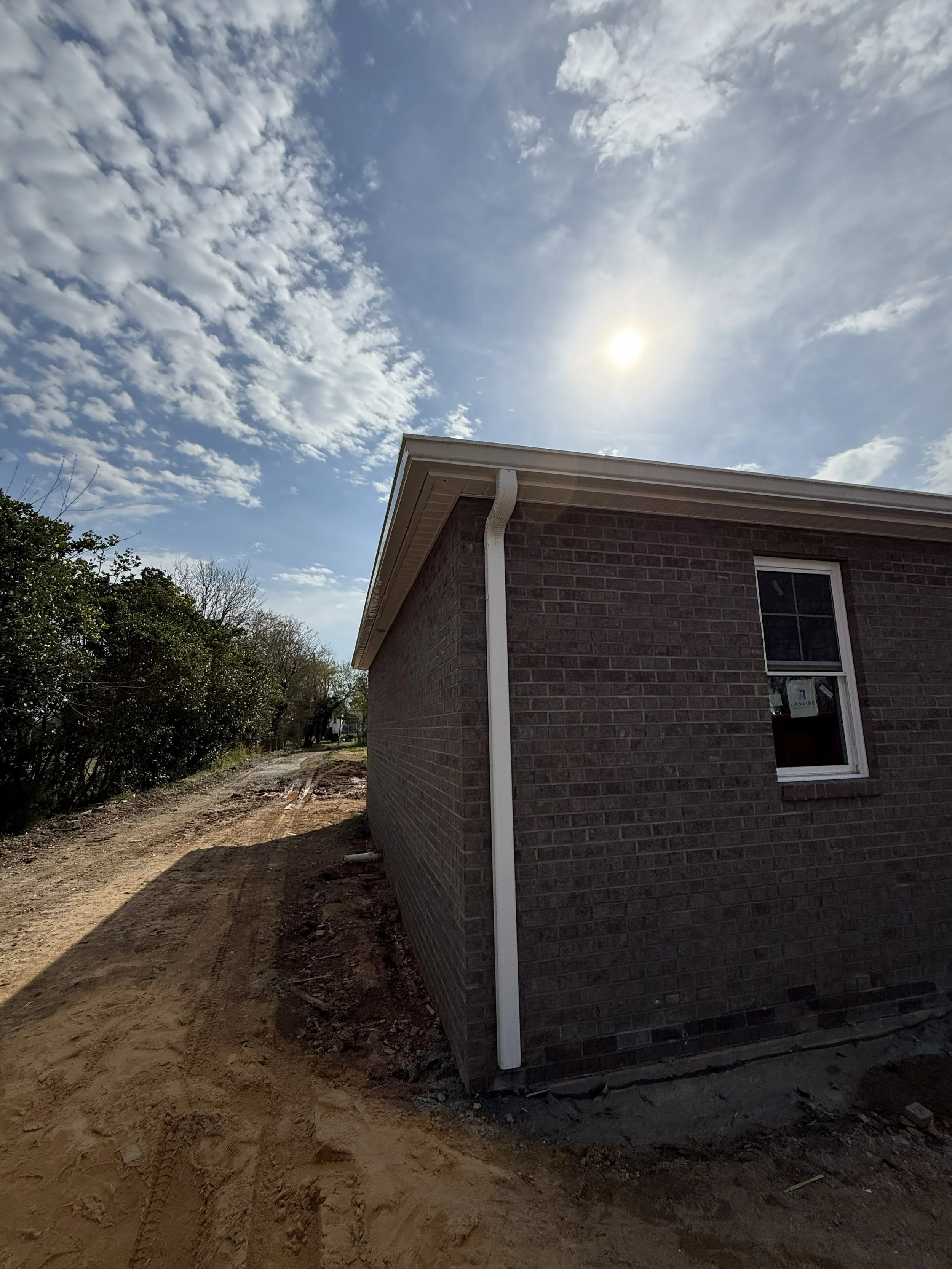 A house with brick exterior wall, white gutter, and window, under a partly cloudy sky with the sun shining, and a dirt road passing by.