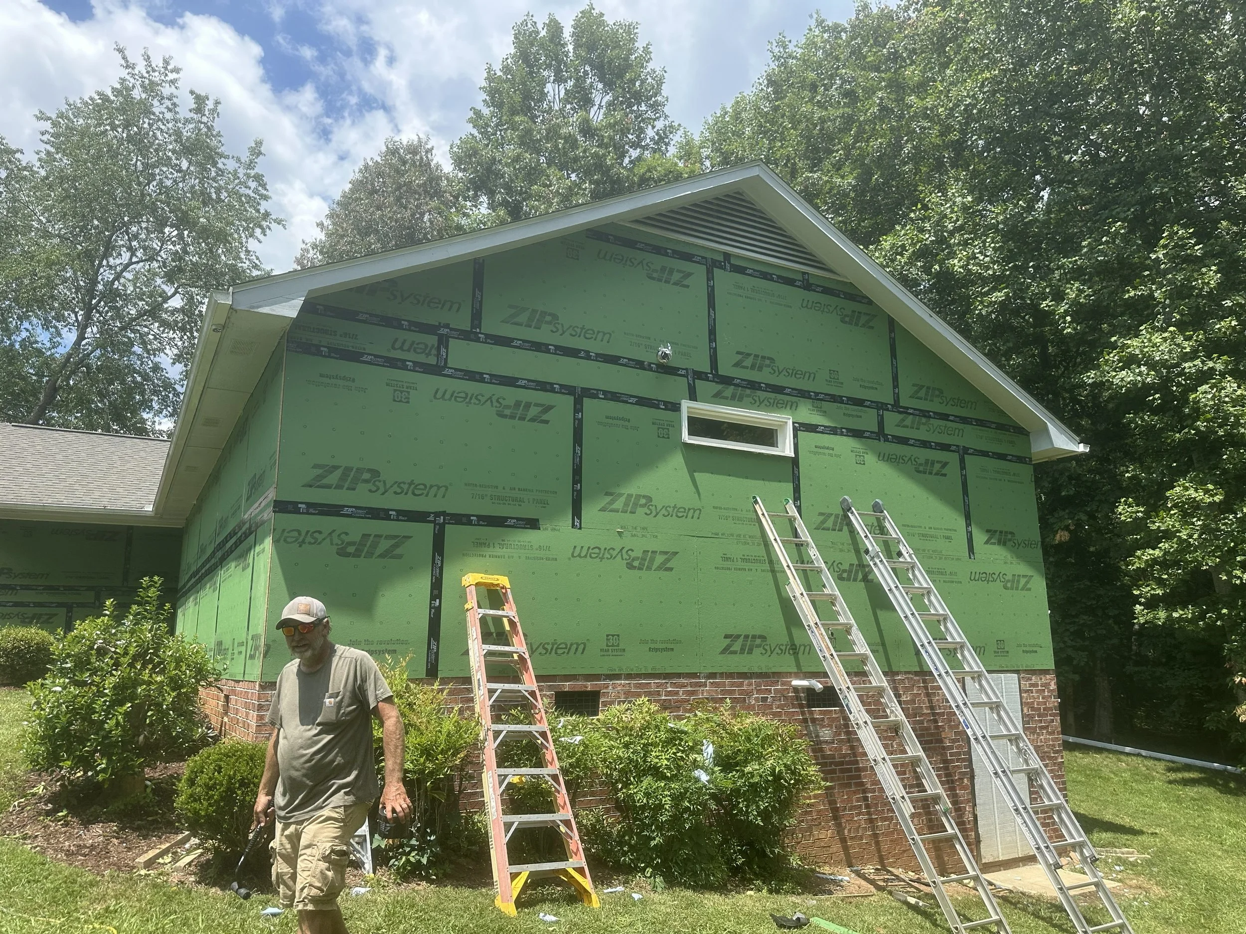 Construction worker in a gray shirt and glasses walking in front of a house under construction, with ladders leaning against the house, and a green house wrap covering the exterior walls, surrounded by trees and bushes.