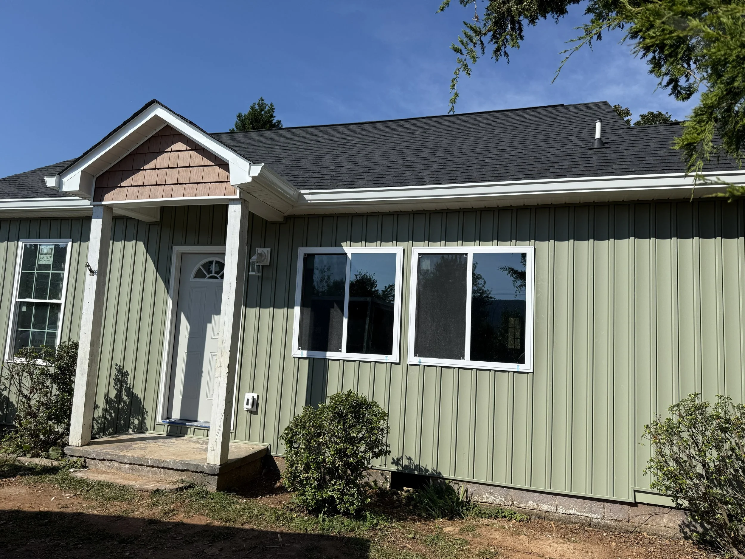The front view of a house with dark gray shingle roof, green siding, and white trim. There is a small porch with white posts and a screen door. Two windows are visible, with one partially covered by a white window screen. Trees and bushes surround th
