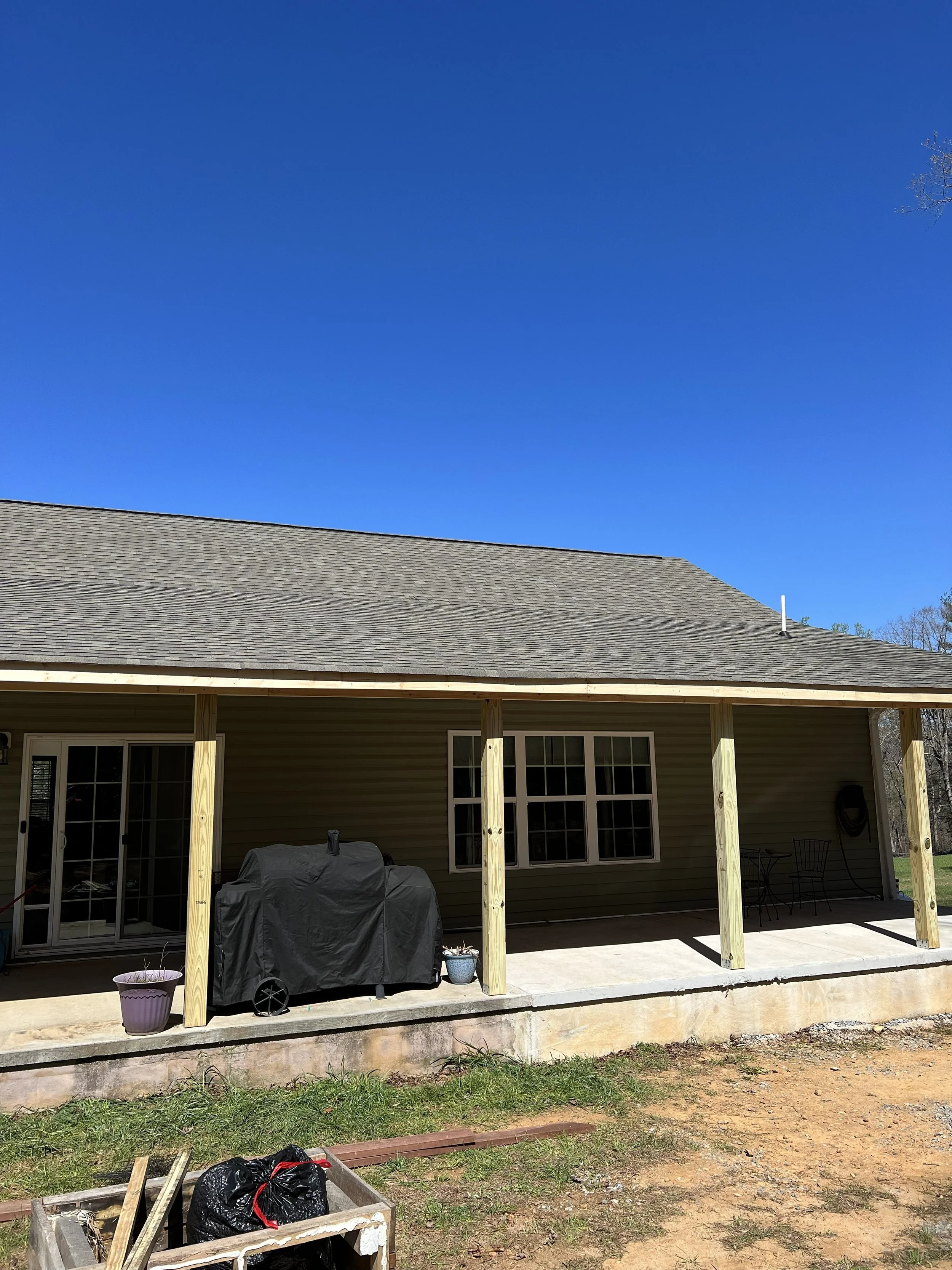 Back porch of a house under construction with wooden posts, roofing, and some outdoor furniture. The yard has some grass, dirt, and construction materials.