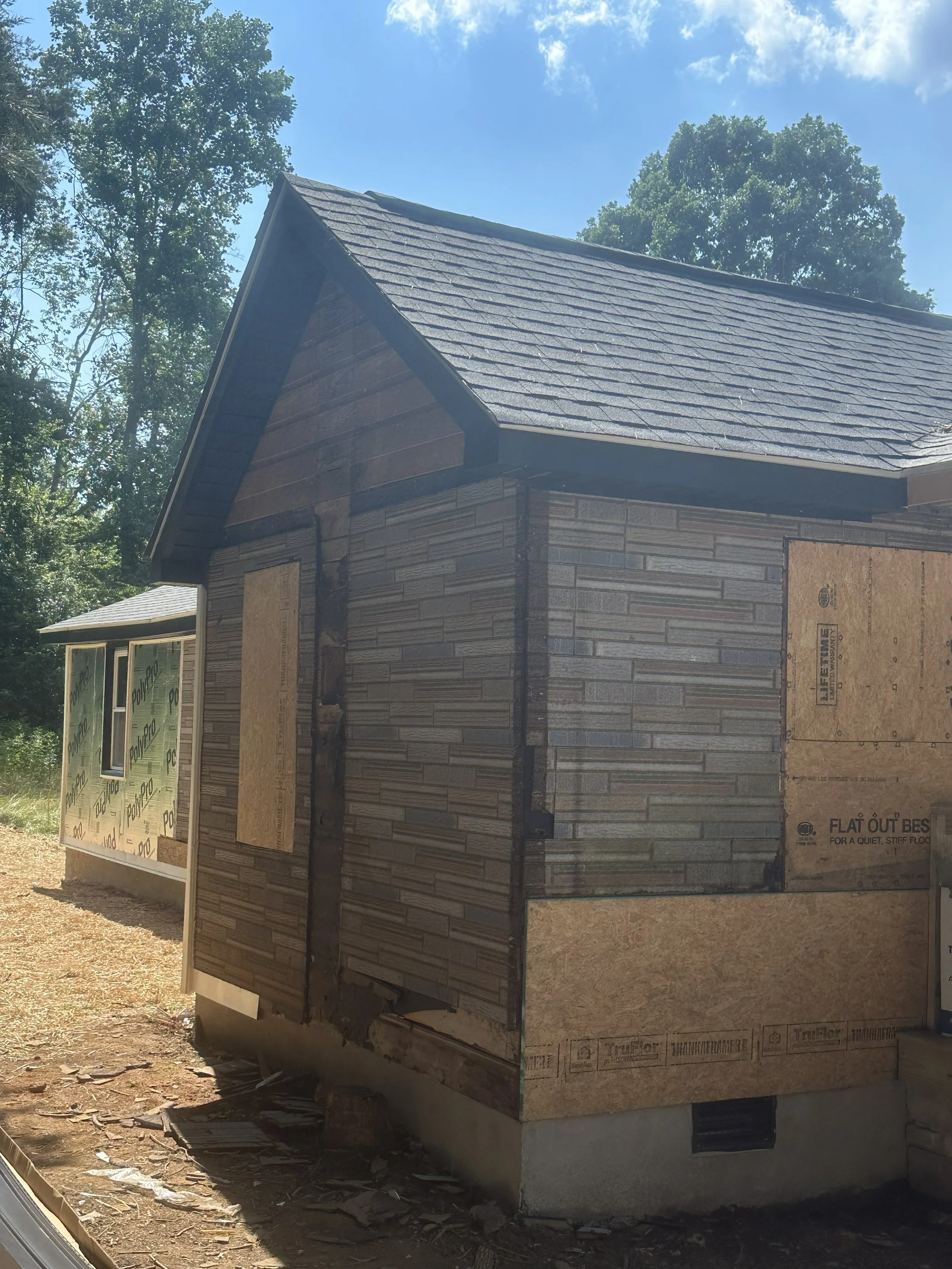 A house under construction with brick siding and plywood on the windows, a peaked roof with black shingles, and surrounded by trees and dirt ground.