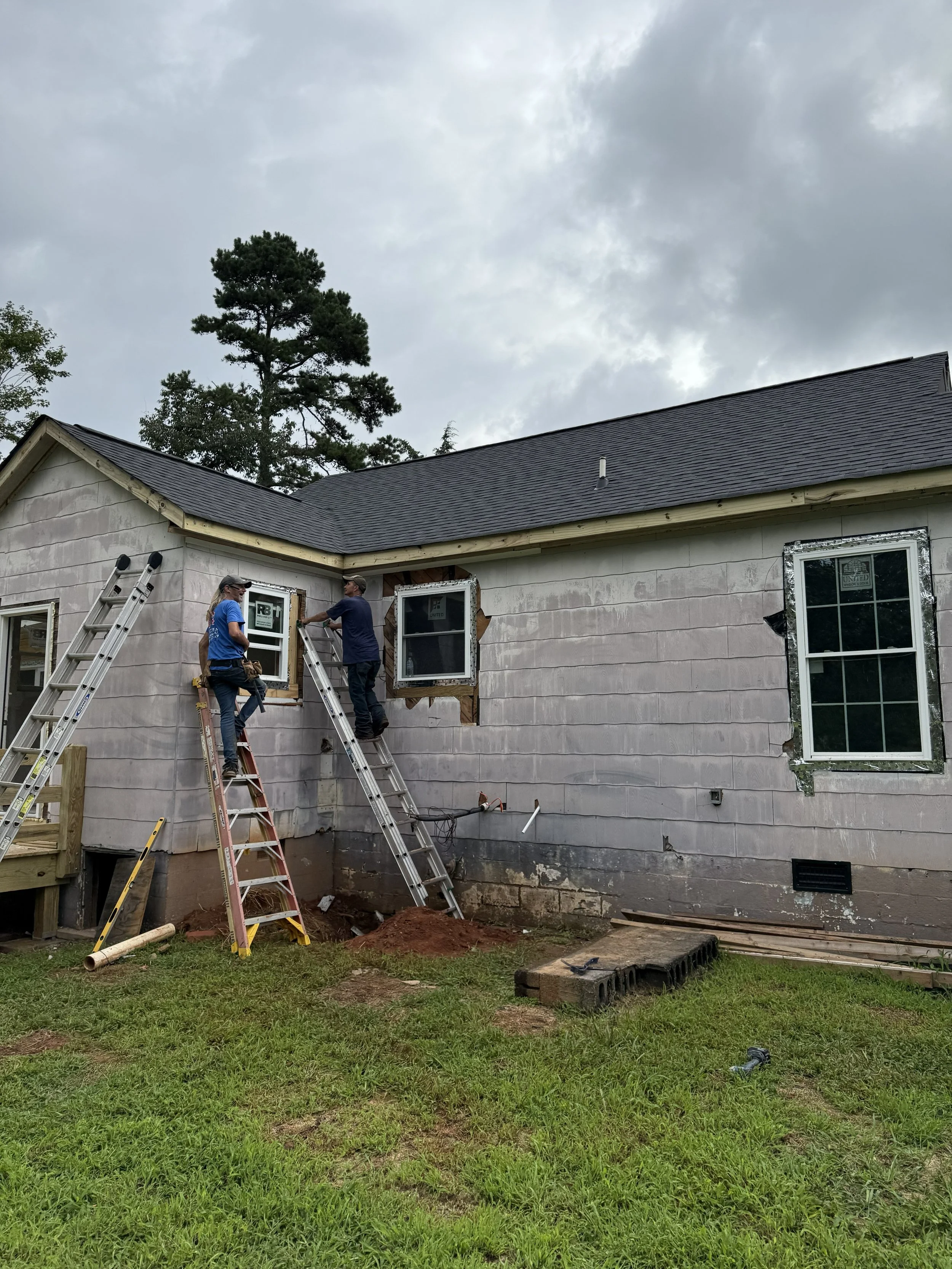 Two construction workers installing a window on the side of a house under construction. The house has an unfinished exterior with visible insulation, and there are ladders, tools, and construction materials on the ground. The sky above is cloudy, and