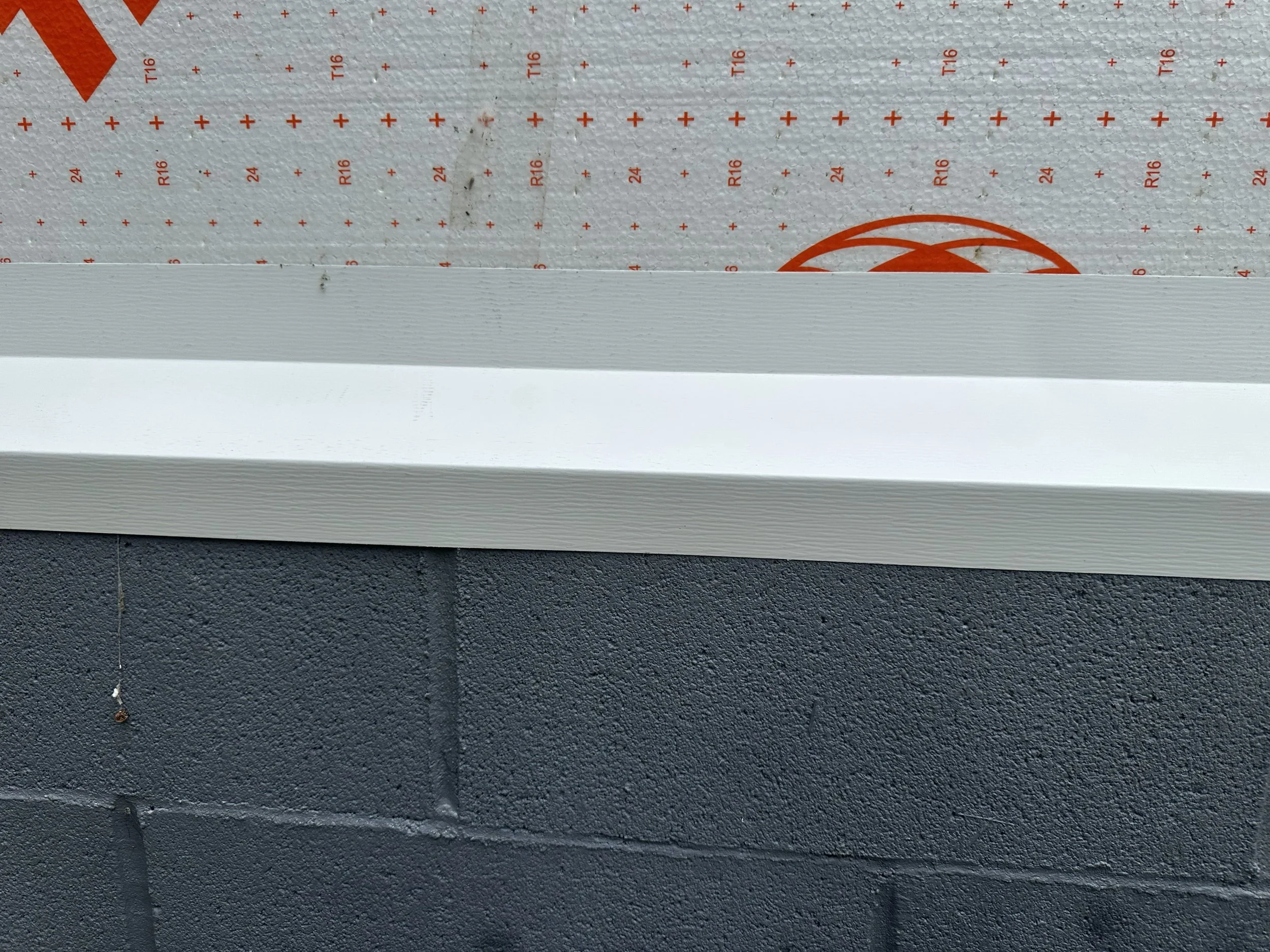 Close-up of a textured white roofing material with orange markings and symbols, installed on a building with a dark-colored wall and some small debris below.