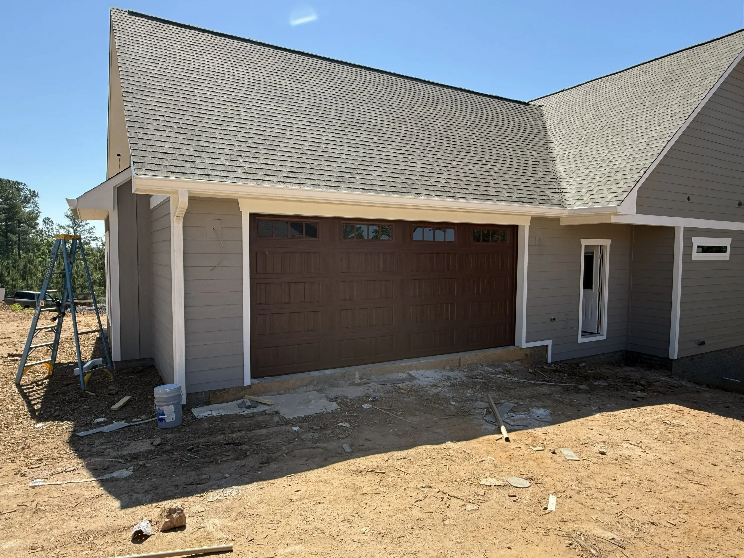 Front of a house under construction with a garage door, partially completed exterior, and building materials and a ladder around the area.