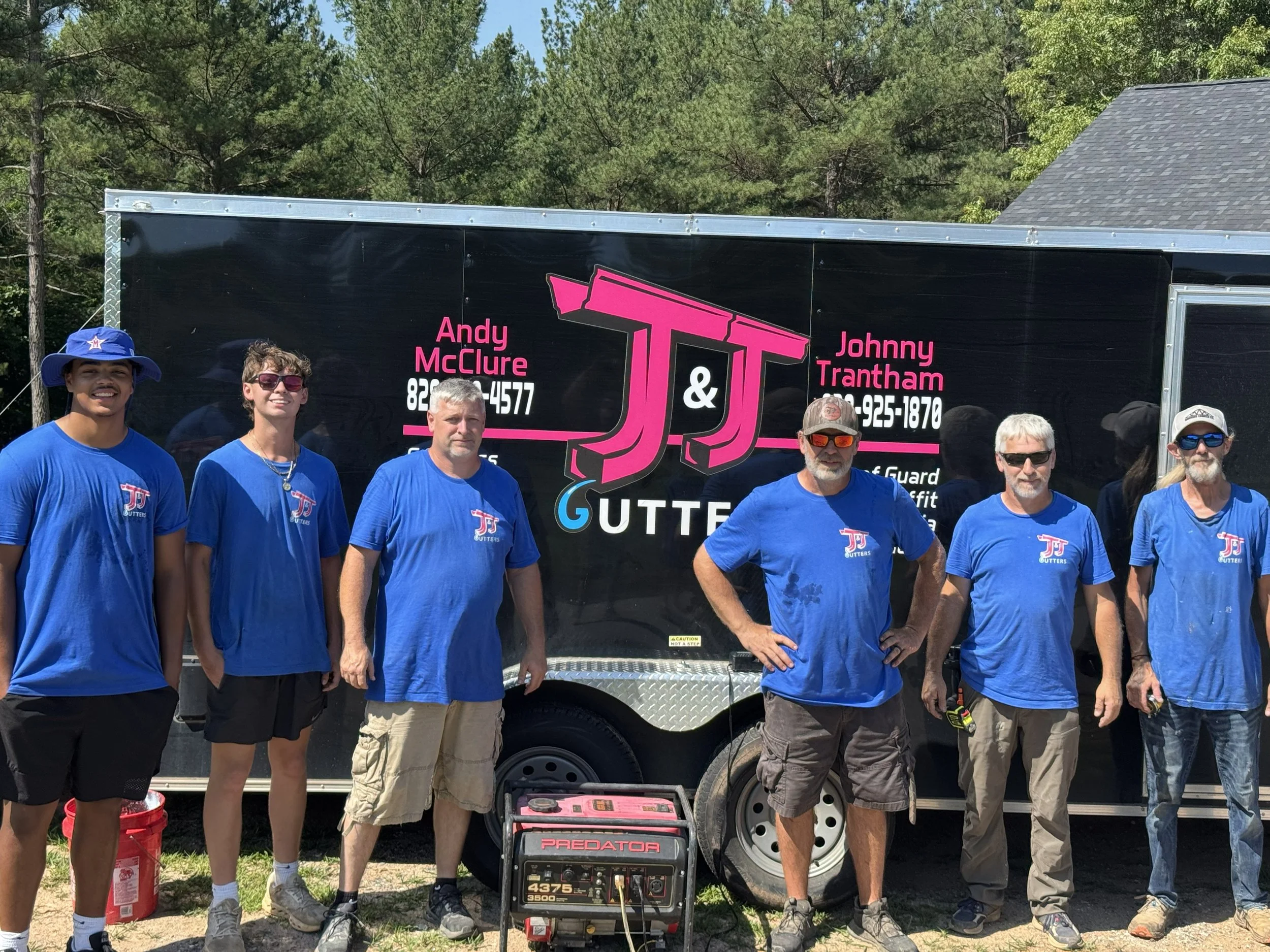 Group of people standing outdoors in front of a trailer with a pink and black logo, wearing matching blue shirts with a logo, some wearing hats and sunglasses, with a generator in front
