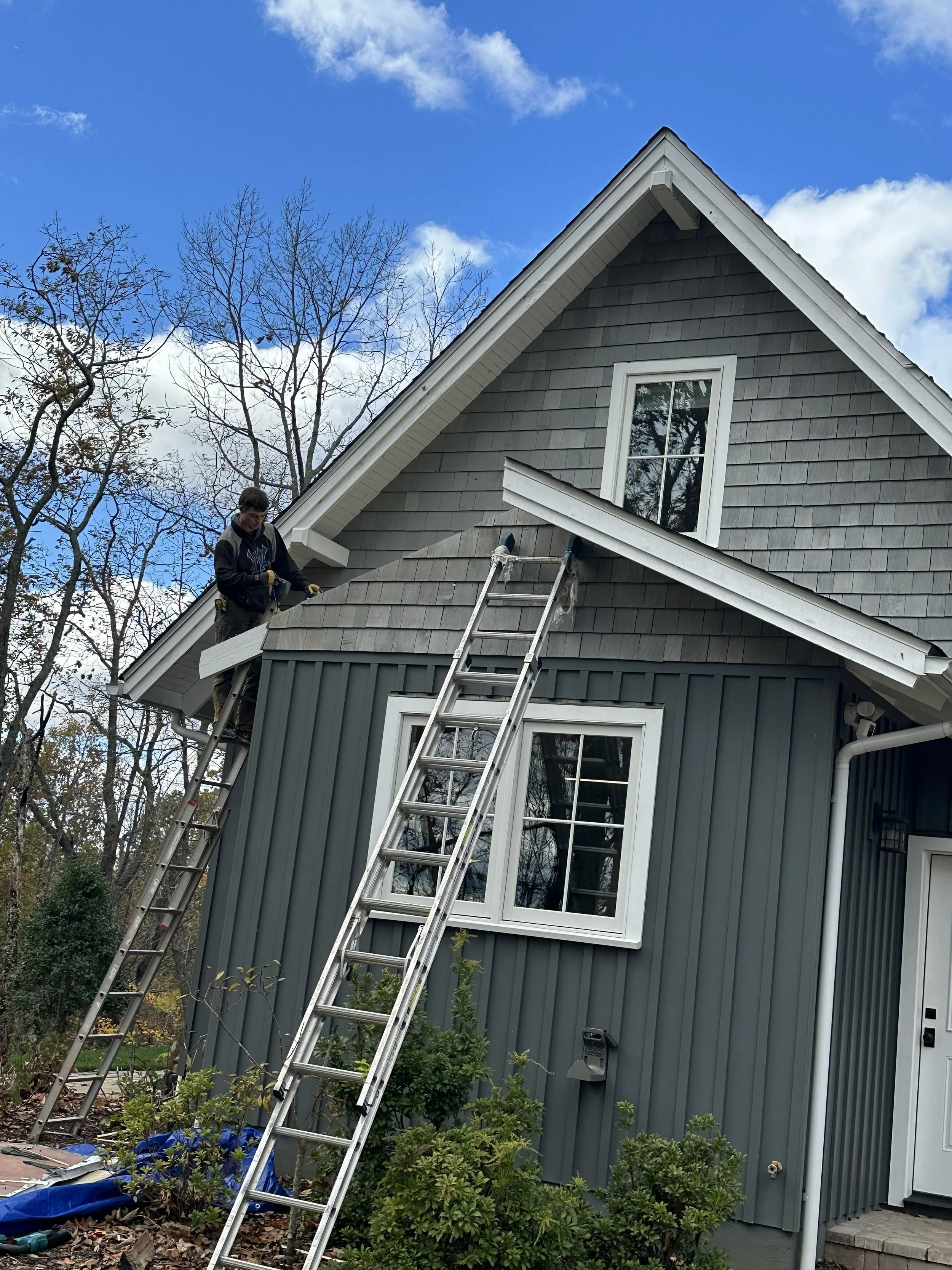 Person working on the roof of a gray house with dark gray shingles, using a ladder for access, surrounded by leafless trees and some bushes, under a partly cloudy sky.