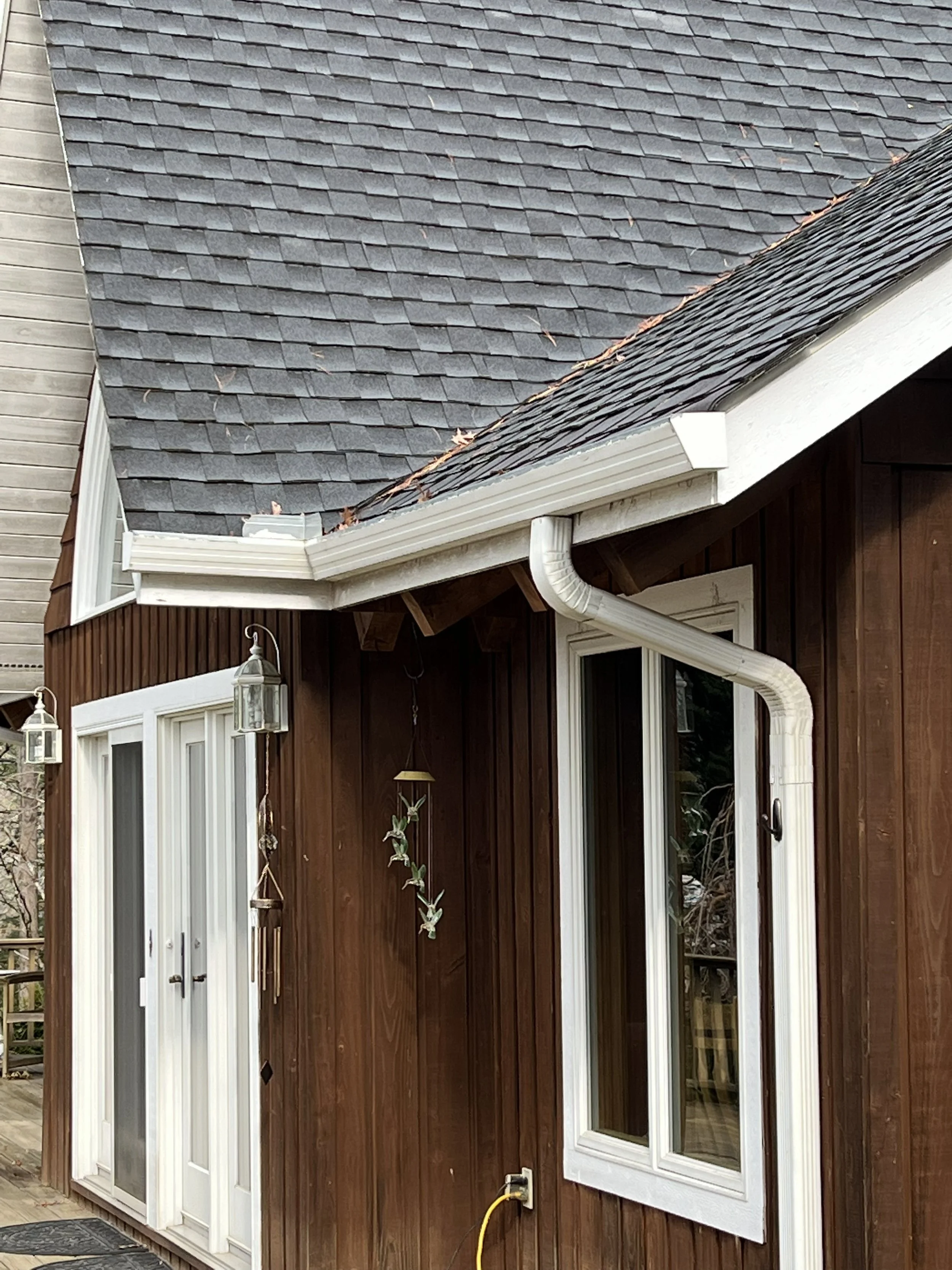 The image shows the exterior of a house with a dark gray shingled roof, white trim, and brown wooden siding. There are two white-framed windows, a white door with glass panels, and an outdoor lantern light fixture. A yellow garden hose is connected t
