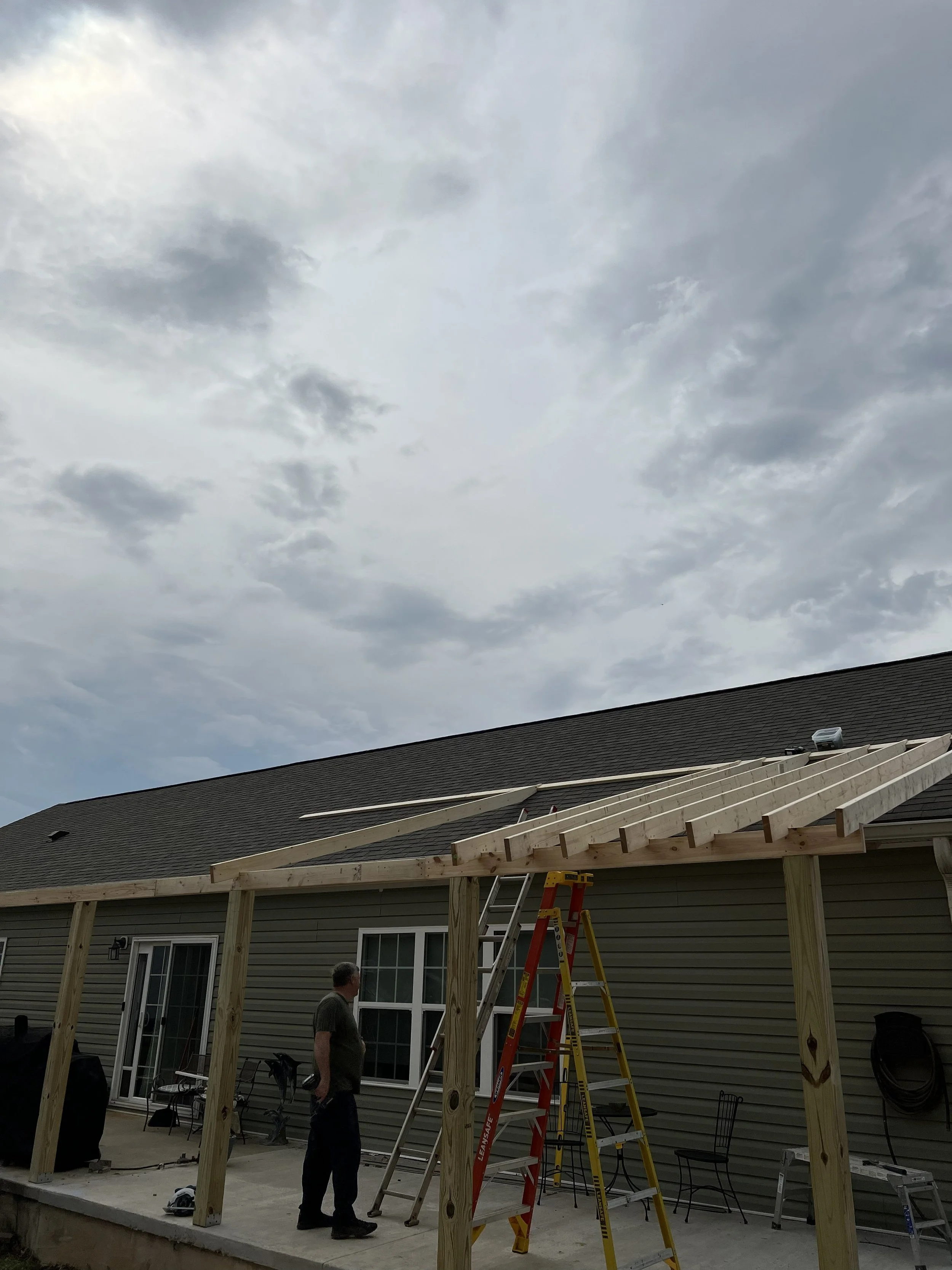 Construction workers building a porch extension on a house with yellow siding, with a ladder and scaffolding, under a cloudy sky.
