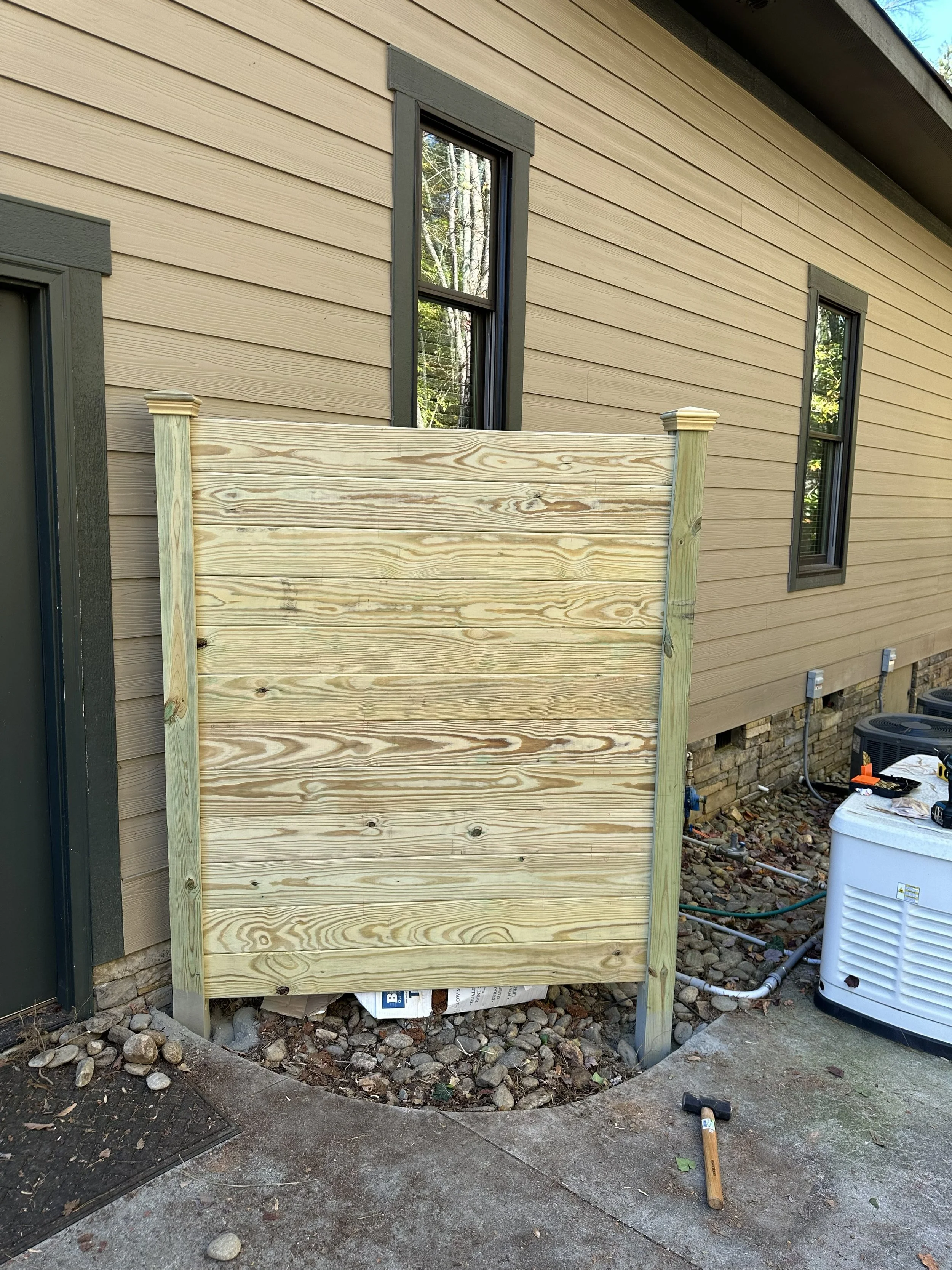 A house exterior with beige horizontal siding and two black-framed windows, and a small unfinished wooden privacy wall or fence under construction, with construction tools and equipment nearby.