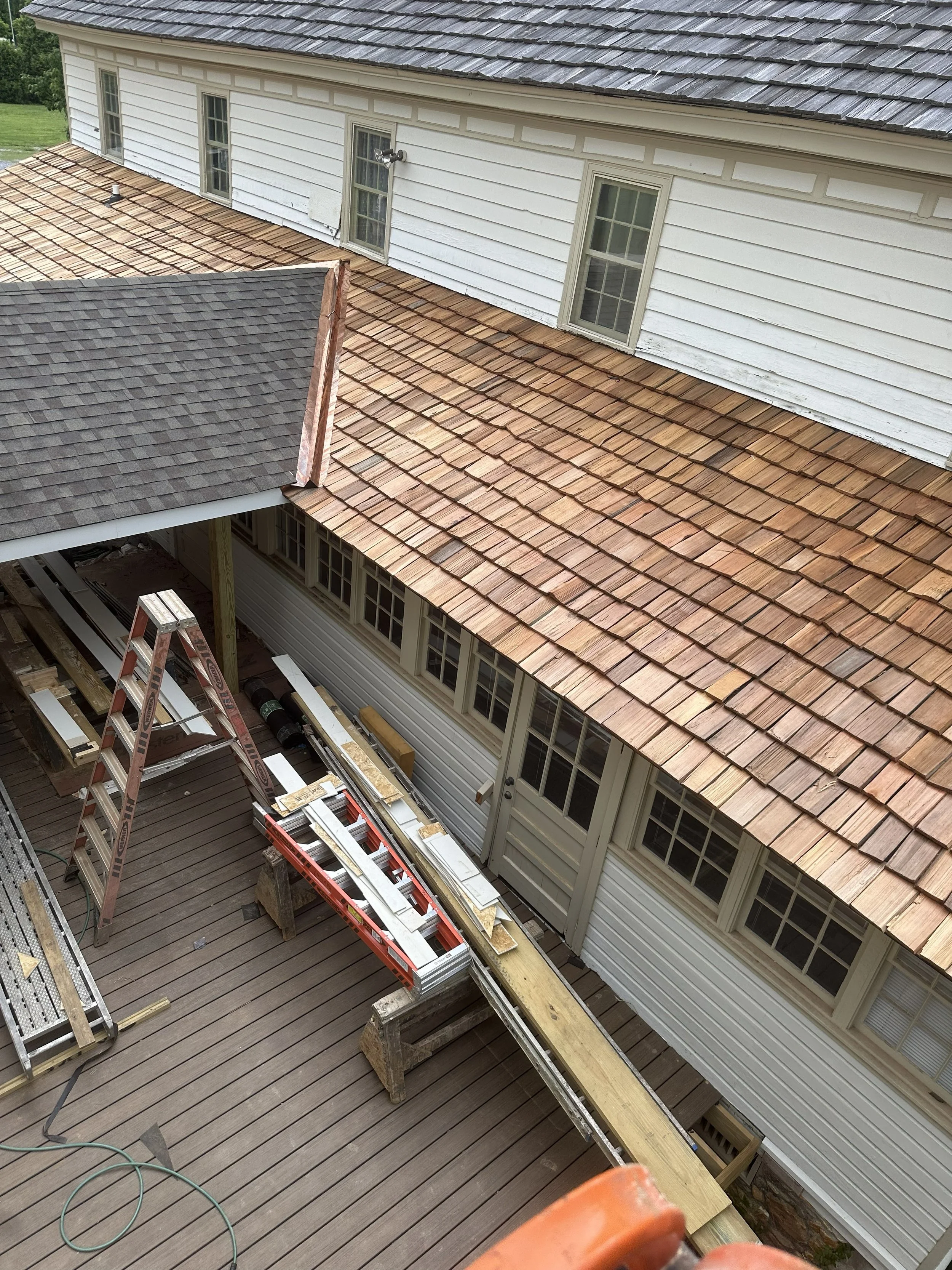 A house with a wooden deck, multiple windows, and a siding exterior under construction. The roof has various shingles, and there are ladders and construction tools on the deck.